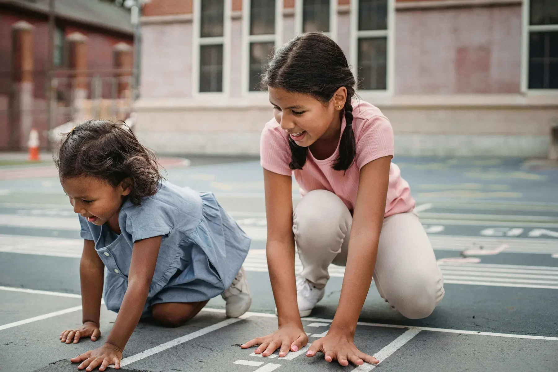 A young girl and a small girl crouching on a running track, preparing to race, outdoors with a school building in the background.