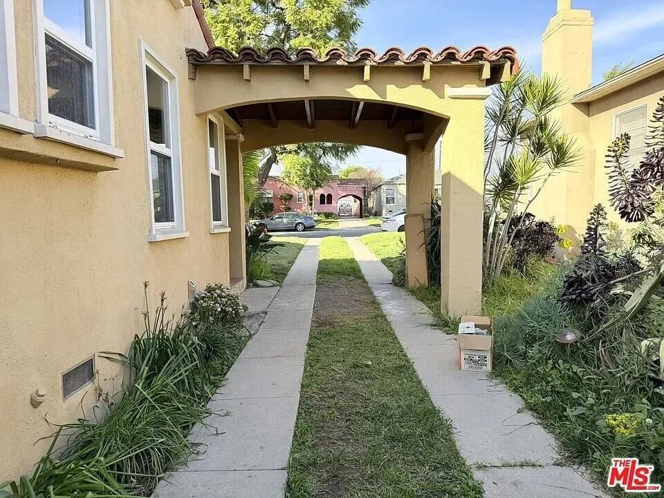 View of a residential area with a yellow stucco building on the left, an archway with a tiled roof, and a grassy walkway leading to a street with parked cars and trees in the background at residence 3016 W 83rd St. in Inglewood California.