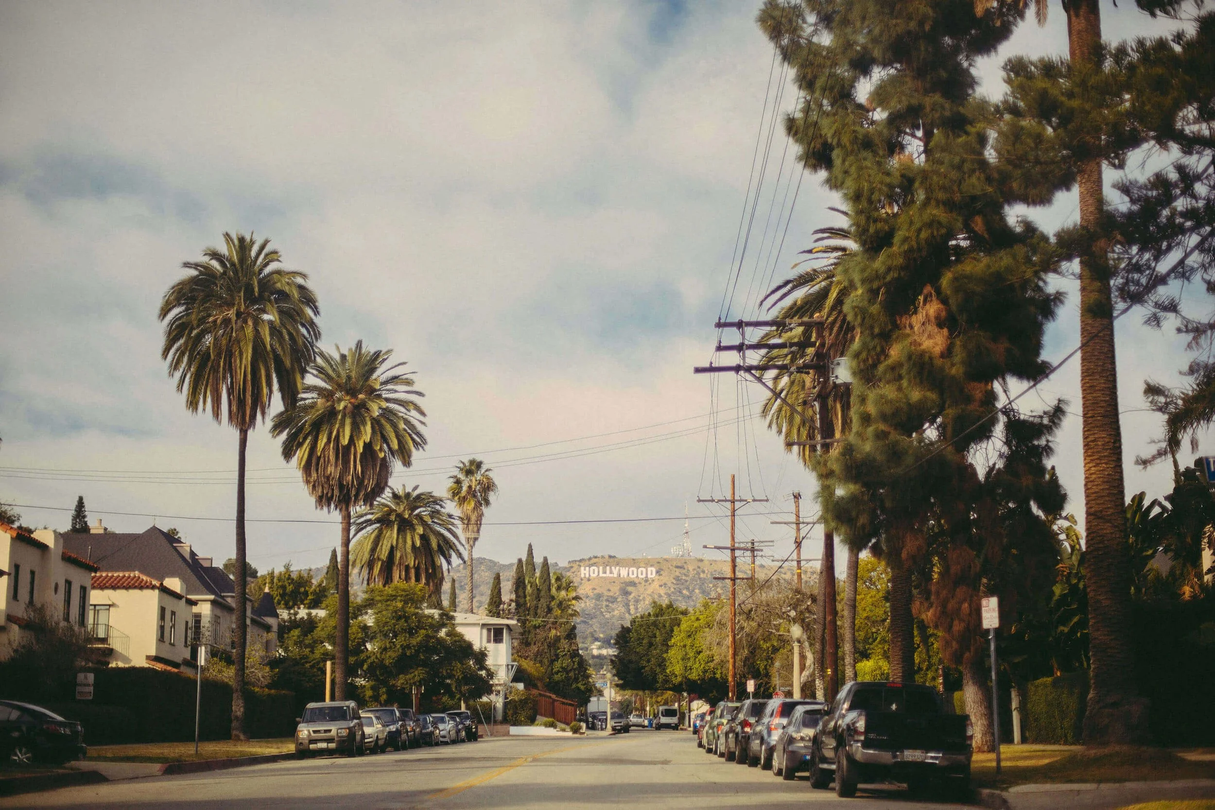 Street view with parked cars, tall palm trees, and the Hollywood sign in the distance on a partly cloudy day.