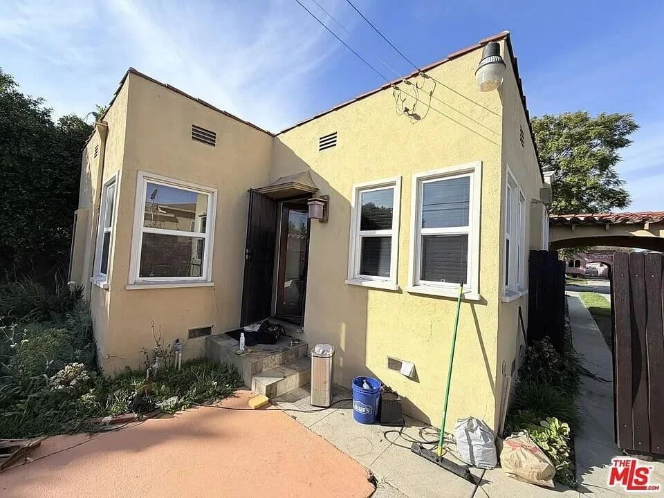 Small, yellow house with four windows, a small covered door entrance, and a few steps leading up to the front door. Items scattered in front of the house include a blue bucket, a trash can, a bag, and a mop. There is a wooden fence to the right and s