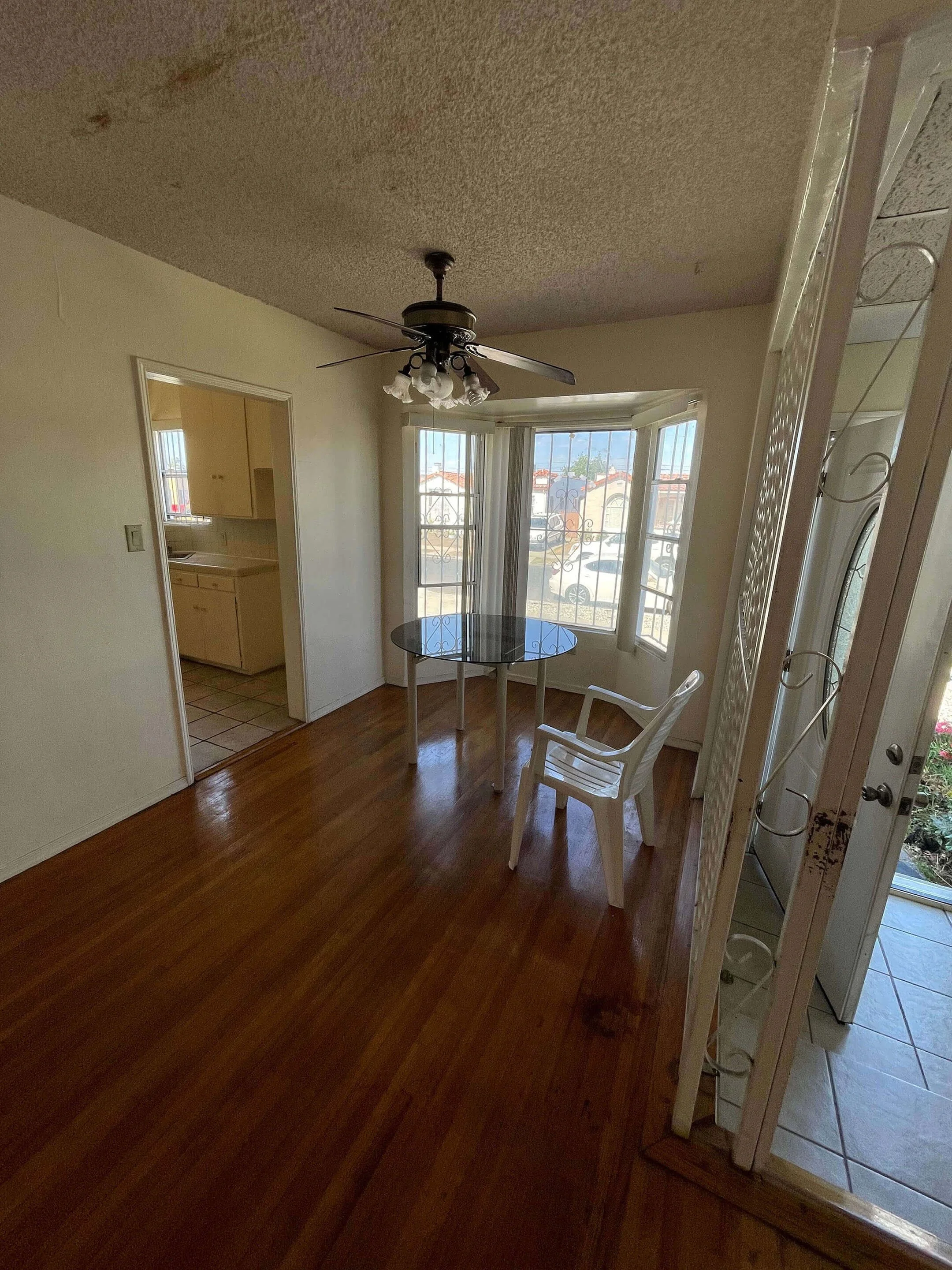 A dining area with a small round glass table, a white plastic chair, a ceiling fan with lights, a bay window with iron bars, and an open doorway to a kitchen. The floor is hardwood, and the adjacent room has tiled flooring.