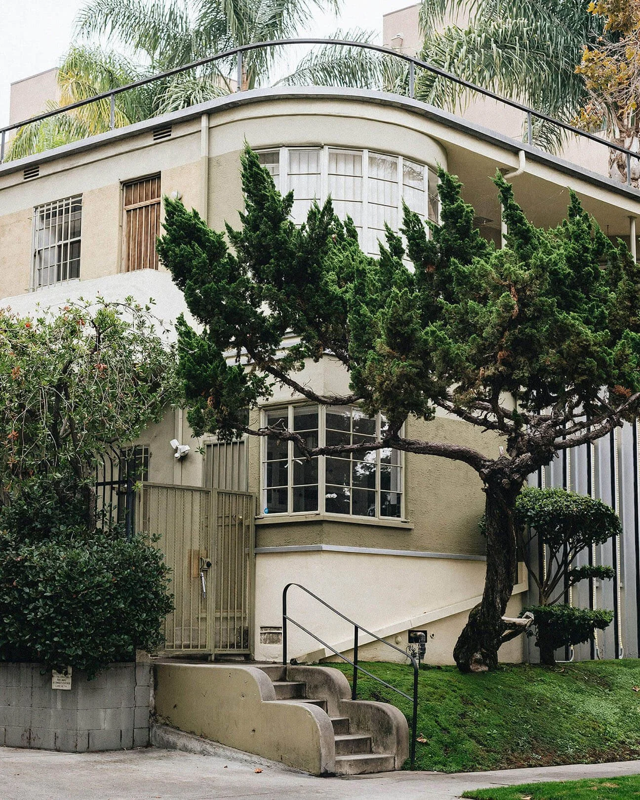 Multi-story building with curved balconies, large windows, and a prominent tree in the front yard with green bushes and stairs leading up to an entrance in Southwest LA.