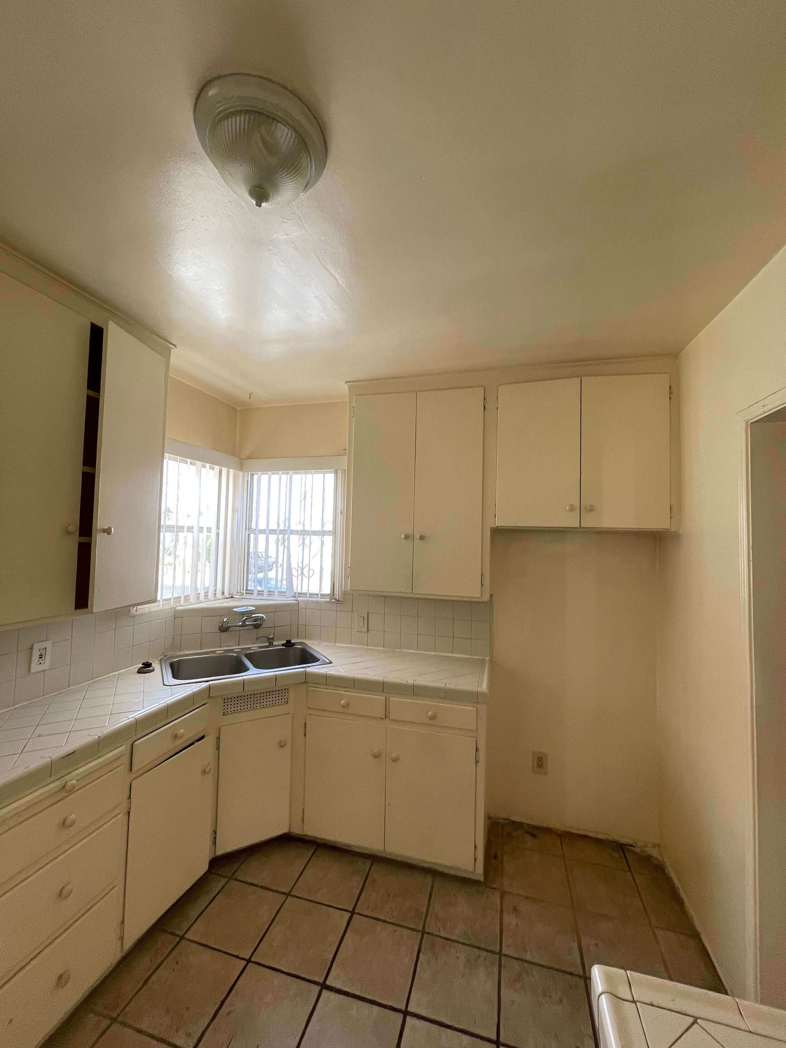 Kitchen at residence 6605 & 6603 4th Ave. with white cabinets, a double sink, a window with vertical blinds, and beige tiled floor. The ceiling has a round light fixture.