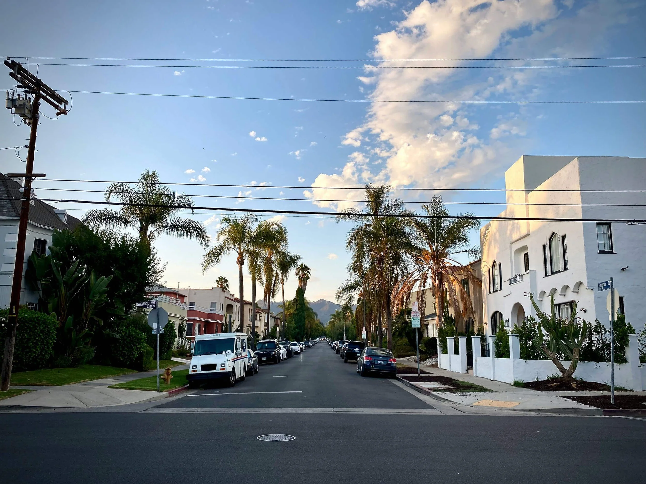A city street scene with parked cars along the curb, a row of palm trees, and residential buildings with white walls. Overhead power lines cross the sky, which is partly cloudy with a few clouds and a blue background.