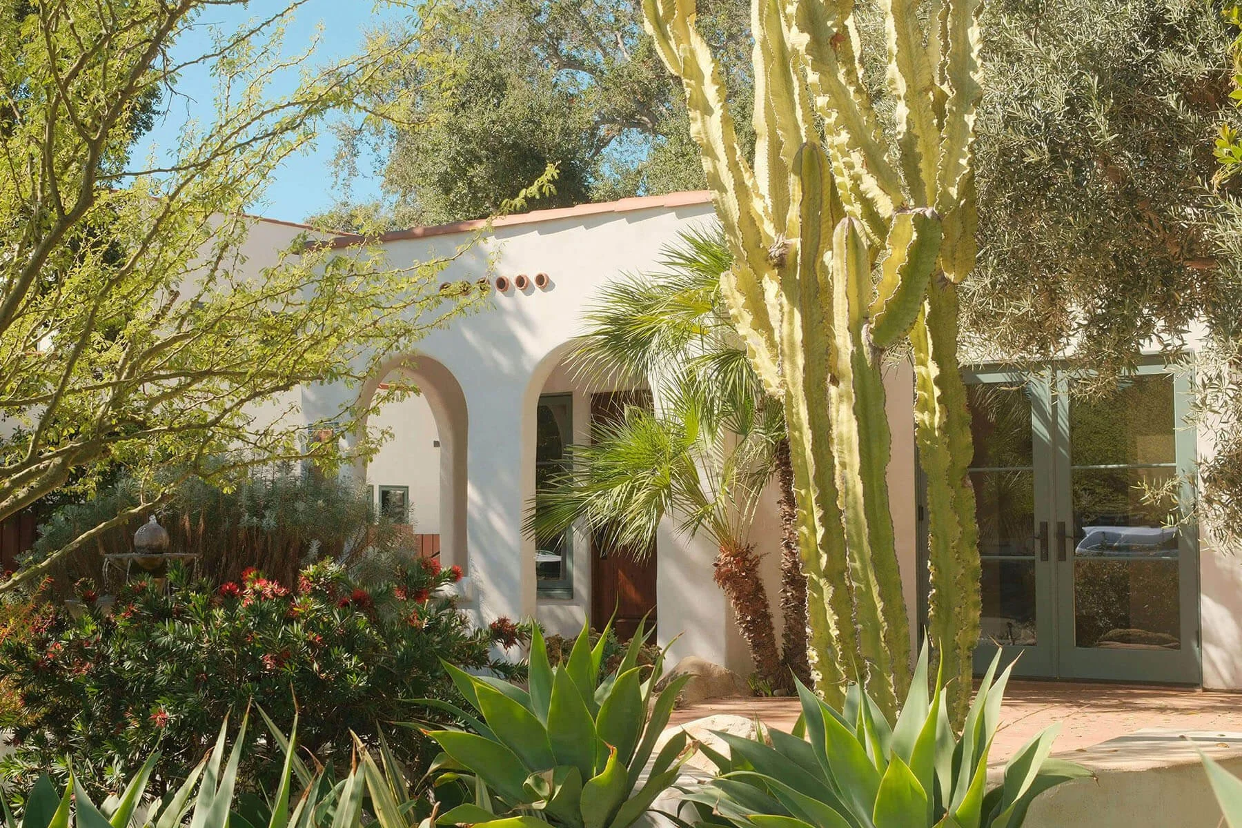 A Spanish-style house with a white stucco exterior, green framed doors, and arched entryways, surrounded by desert plants including a tall cactus and various shrubs and trees, under a clear blue sky.