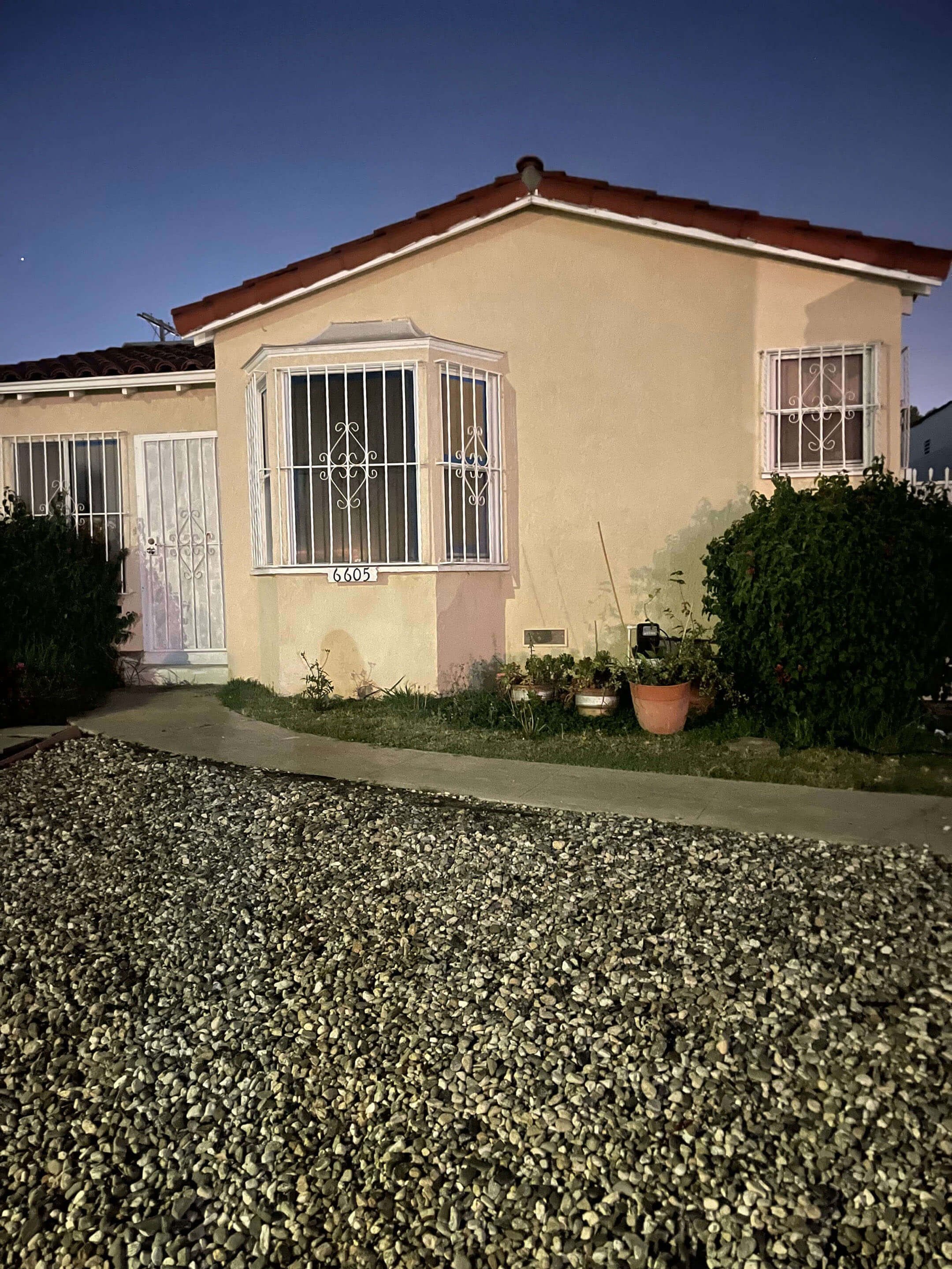 Front view of a beige house with barred windows and a red-tiled roof, night scene with a star visible in the sky, front yard with pots of plants, and a gravel pathway.