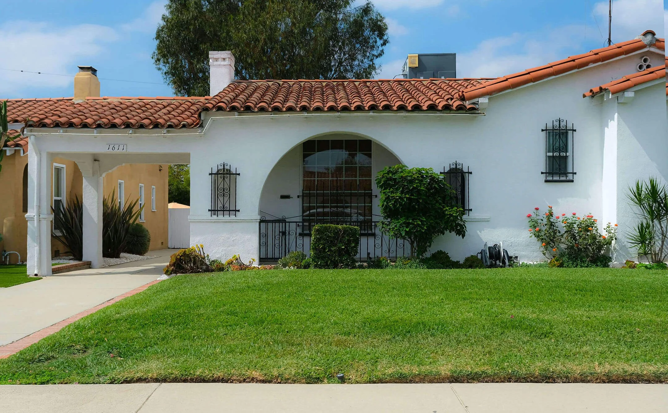 Front view of a Spanish-style white house with a red tile roof, small front yard with green grass, and decorative black window grills. There are flowering bushes and plants near the house, a sidewalk in the foreground, and the sky with clouds above.