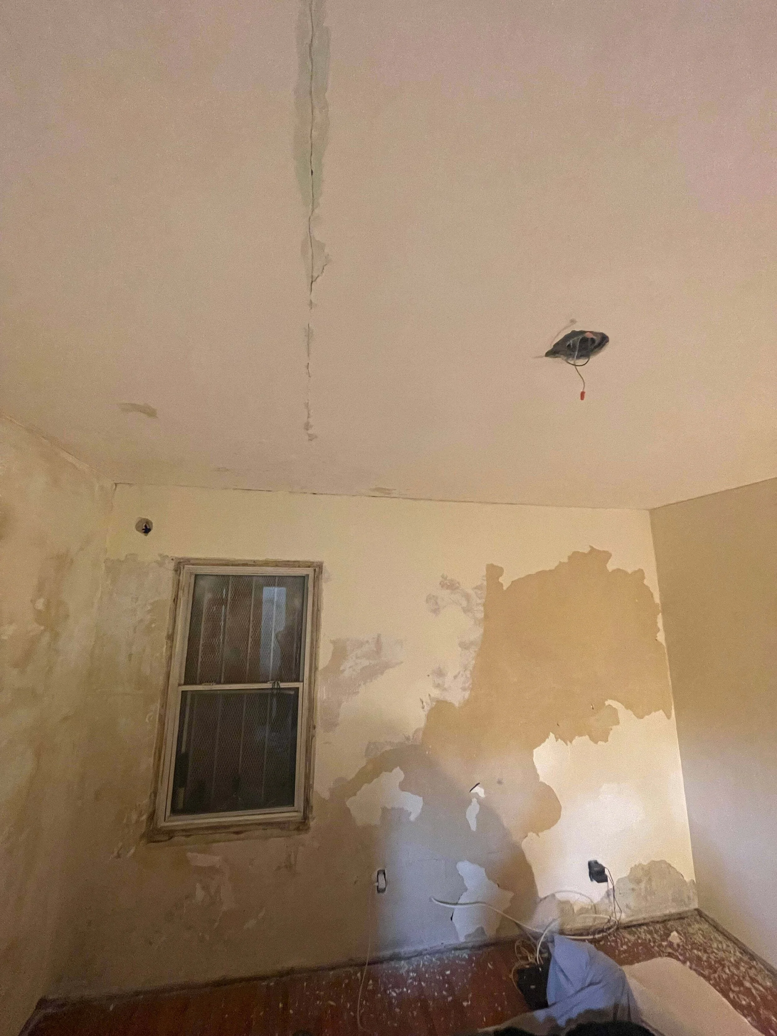 6605 & 6603 4tth Ave. under renovation with peeling paint, exposed wires, and a damaged ceiling with a large crack running down. A window with a wooden frame is on the wall. The floor has debris, and there is a bag and some tools on the floor.