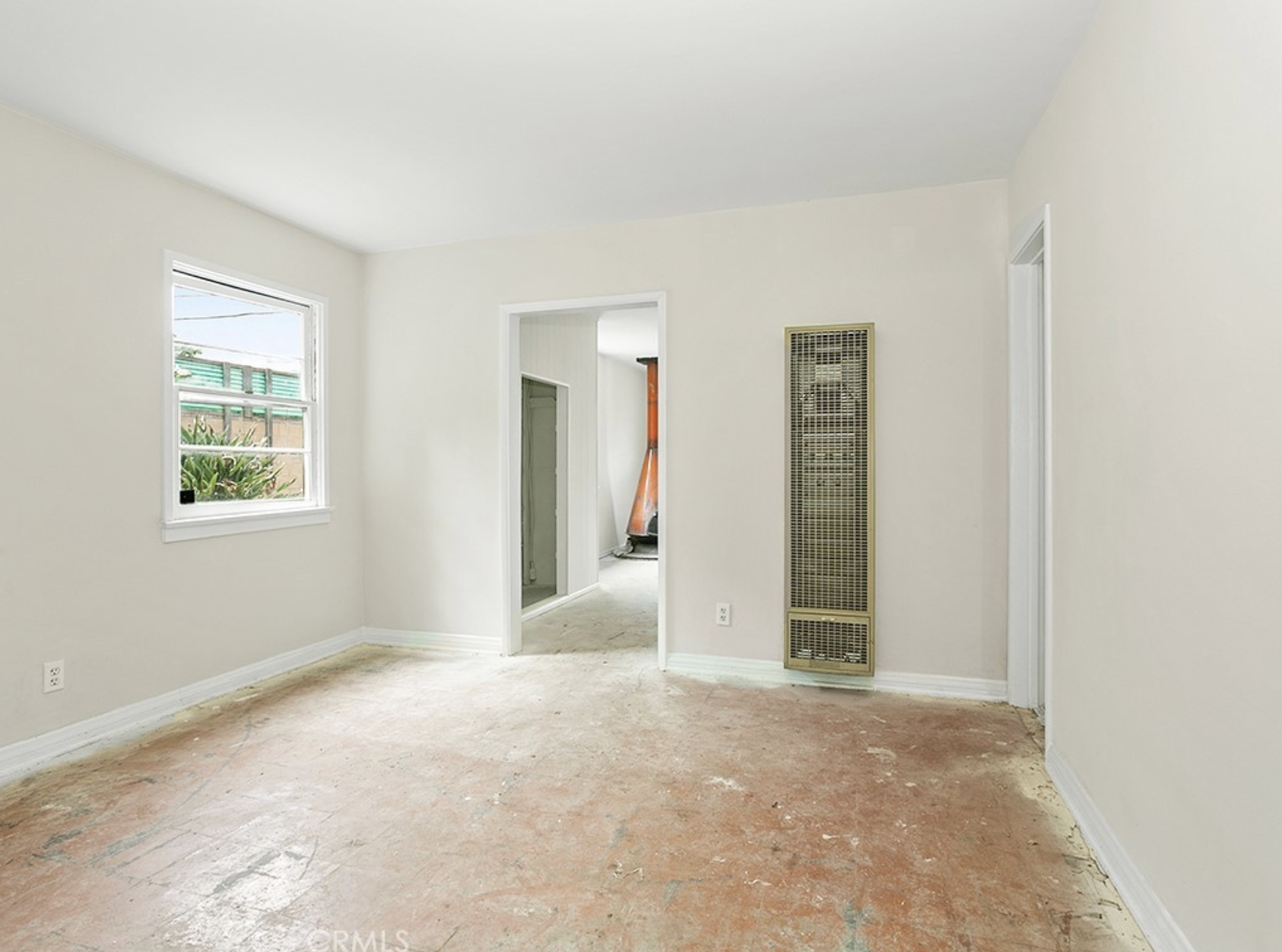 Empty room with white walls, a window with outside plants, an open doorway, a wall heater, and a beige carpeted floor at residence 5645 & 5645 ½ Auckland Ave, in North Hollywood, California.
