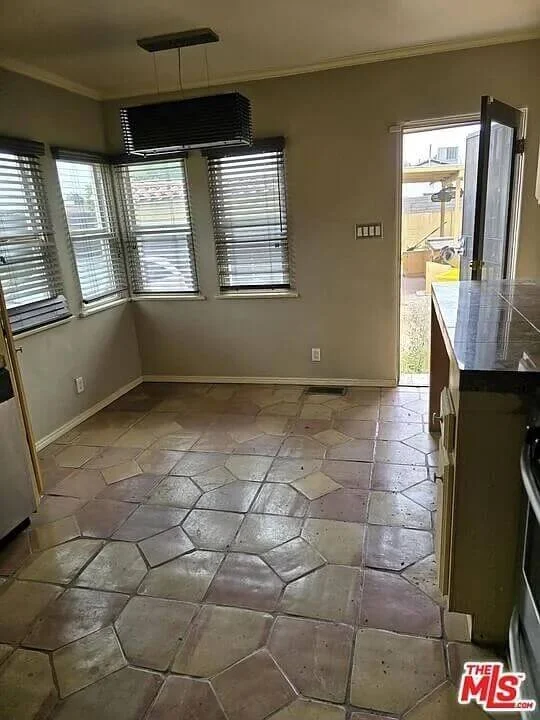 Empty kitchen area with beige walls, large windows with blinds, and a door leading outside. The floor has large, hexagonal, beige tiles at residence 3016 W 83rd St. in Inglewood California.