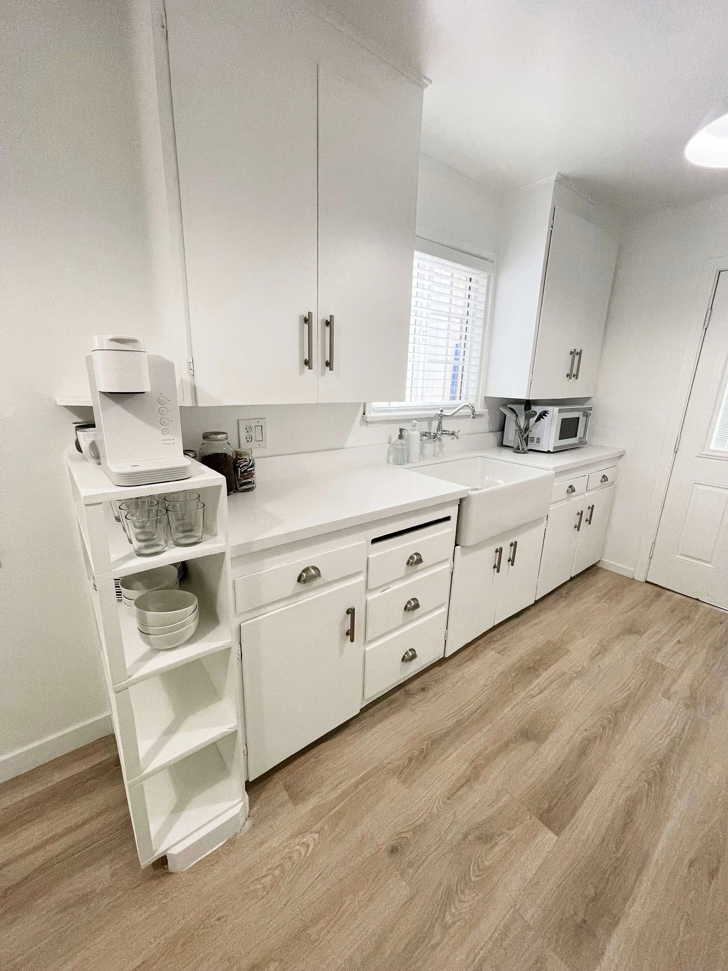 A white kitchen with white cabinets, a microwave, a coffee maker, and a small white shelf with glasses and bowls. There is a window above the sink and wood flooring.