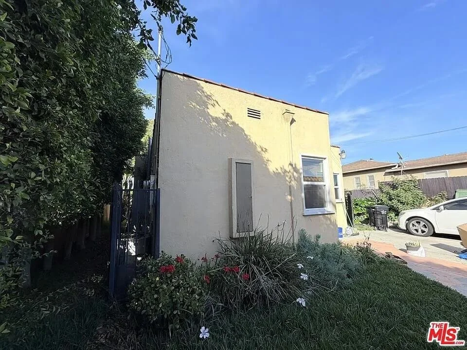Side view of a small, beige stucco building with a window, surrounded by plants and a garden, with neighboring houses and cars in the background under a clear blue sky at residence 3016 W 83rd St. in Inglewood California.