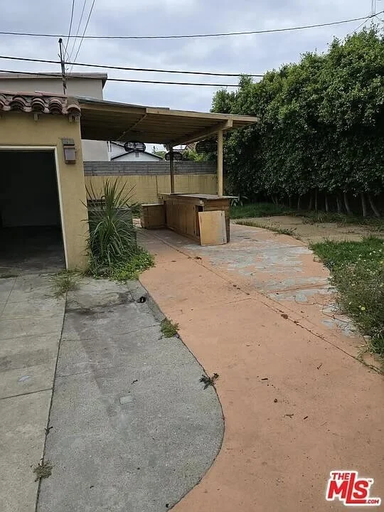 Backyard with a concrete and asphalt pathway, a covered patio area with wooden tables, surrounded by green bushes and trees, under a cloudy sky at residence 3016 W 83rd St. in Inglewood California.