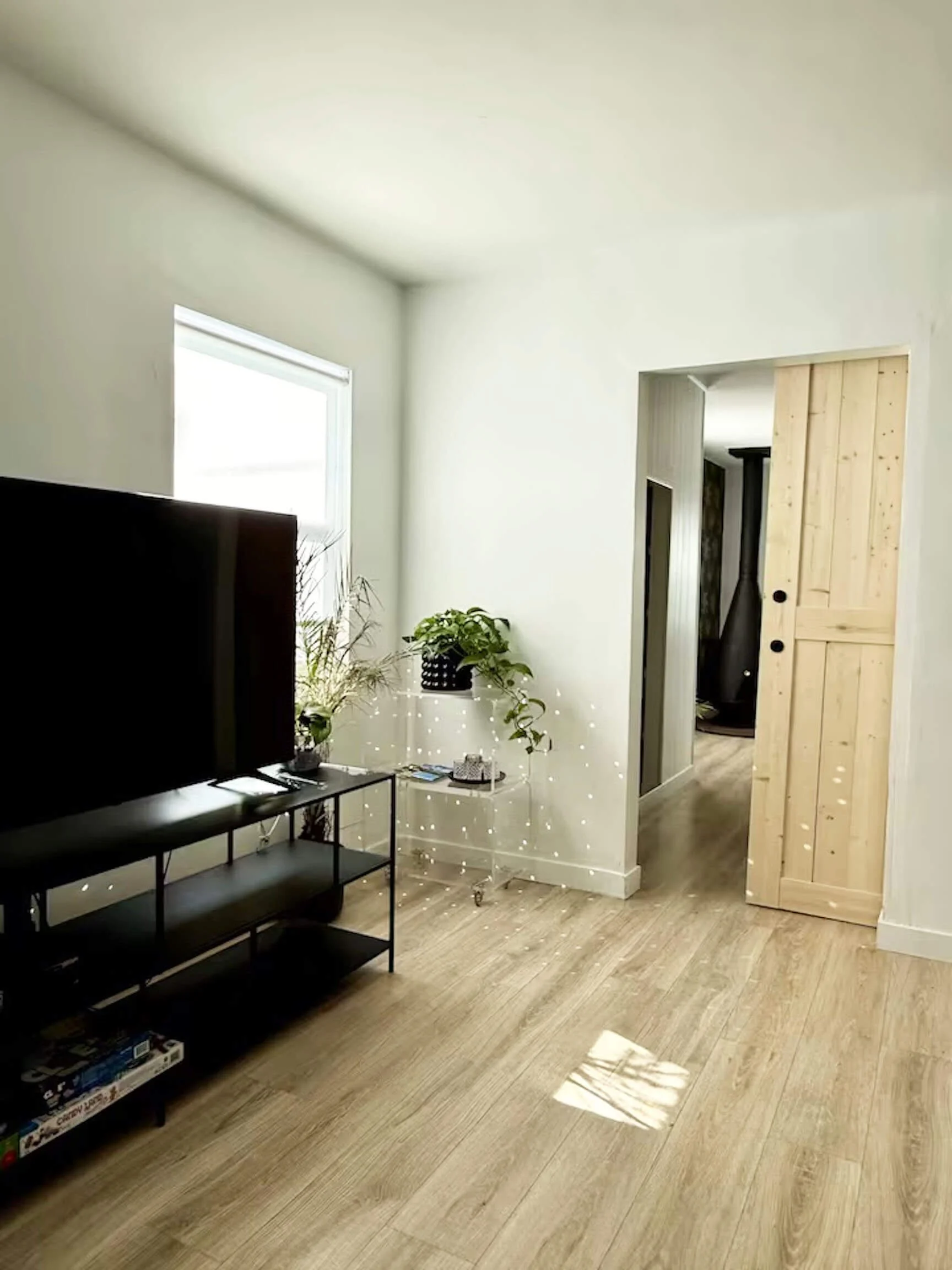 Living room with a black TV stand, a large flat-screen TV, potted plants, and a wooden sliding door, with light-colored walls, a window, and wooden flooring.