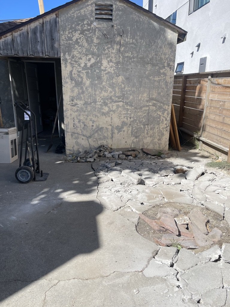 A small outdoor area with an old, weathered concrete building on the left, a wooden fence on the right, and broken cracked concrete pavement with a circular stone platform in the ground at residence 5645 & 5645 ½ Auckland Ave, Hollywood California.
