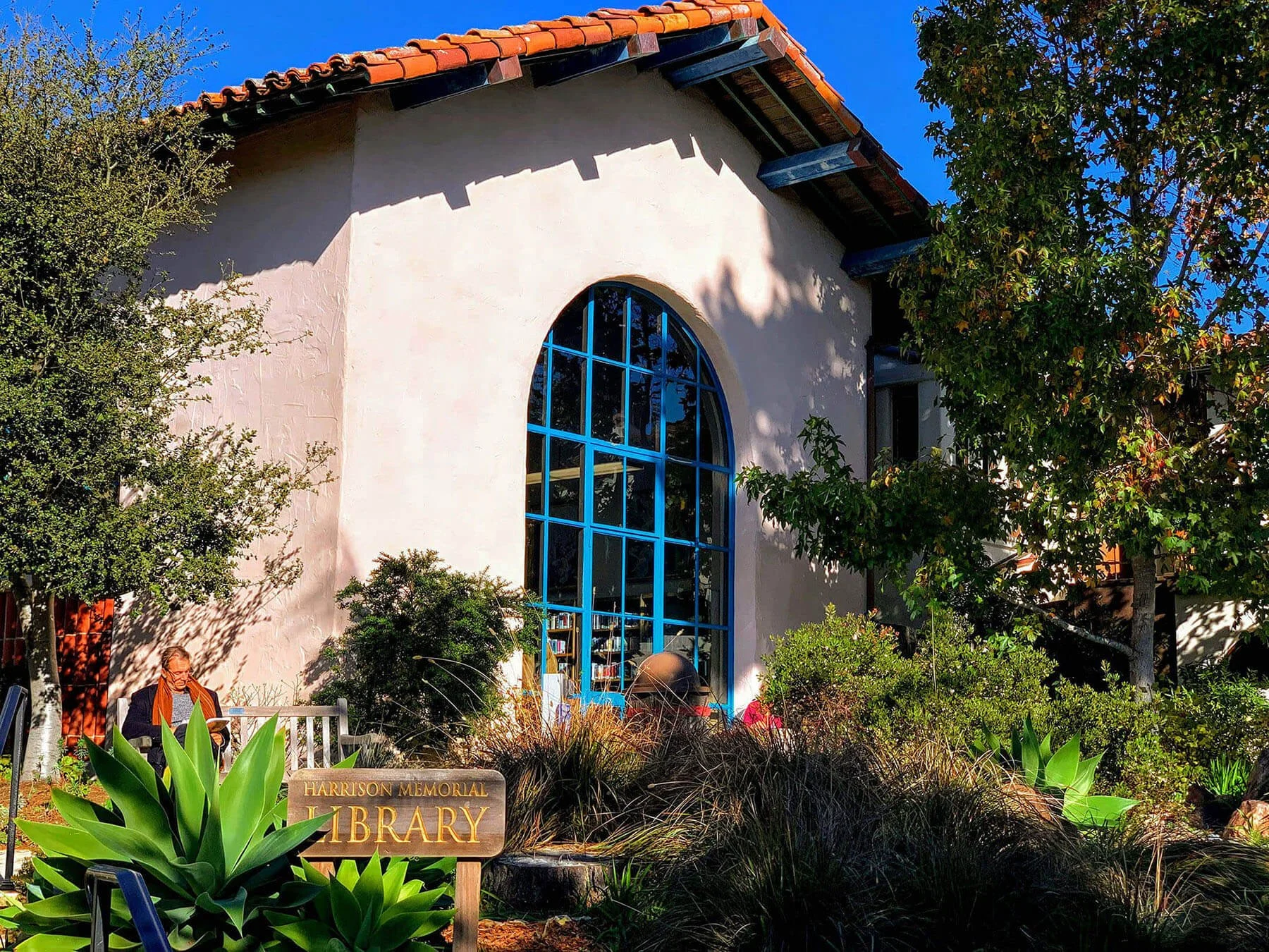 Exterior view of the Harrison Memorial Library with a large arched blue window, surrounded by green plants and trees. A person is sitting on a white bench, and there is a wooden sign in the foreground that reads 'Harrison Memorial Library'. The building has a tiled roof and a white stucco wall, with clear blue sky visible above.