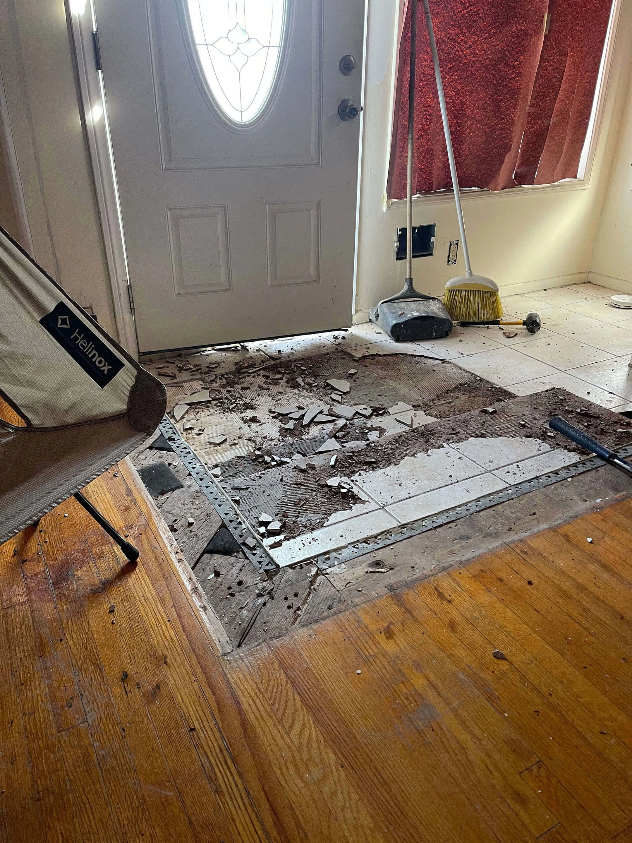 A household entrance with a white door, partially opened, revealing a cloudy day outside. The floor in front of the door shows damage with broken tiles and debris scattered across, with some tiles removed to reveal the subfloor. There are cleaning to