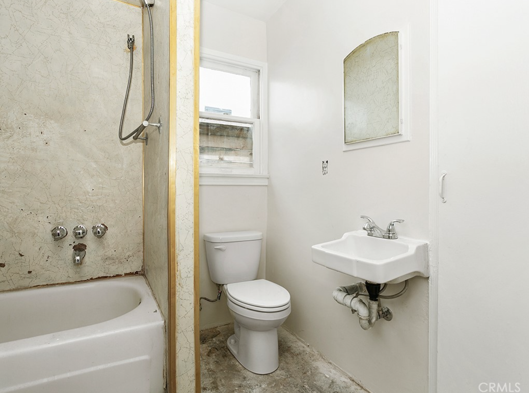 A small bathroom with a toilet, a small sink with a mirror above, and a shower with a partial door. The shower has a worn wall with three knobs and a showerhead hanging from a flexible hose. The floor appears unfinished or dirty, and there's a window