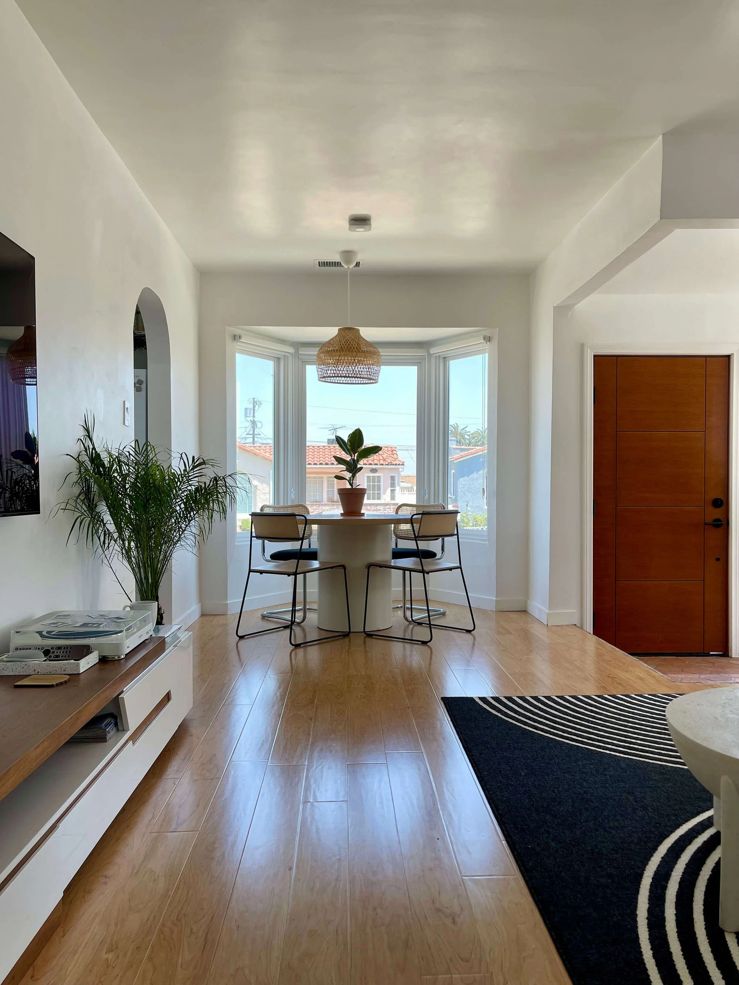 Bright dining area with large bay window, round table, and four chairs, decorated with a potted plant and woven pendant lamp. Walls painted white, hardwood flooring, and an entry door to the right.