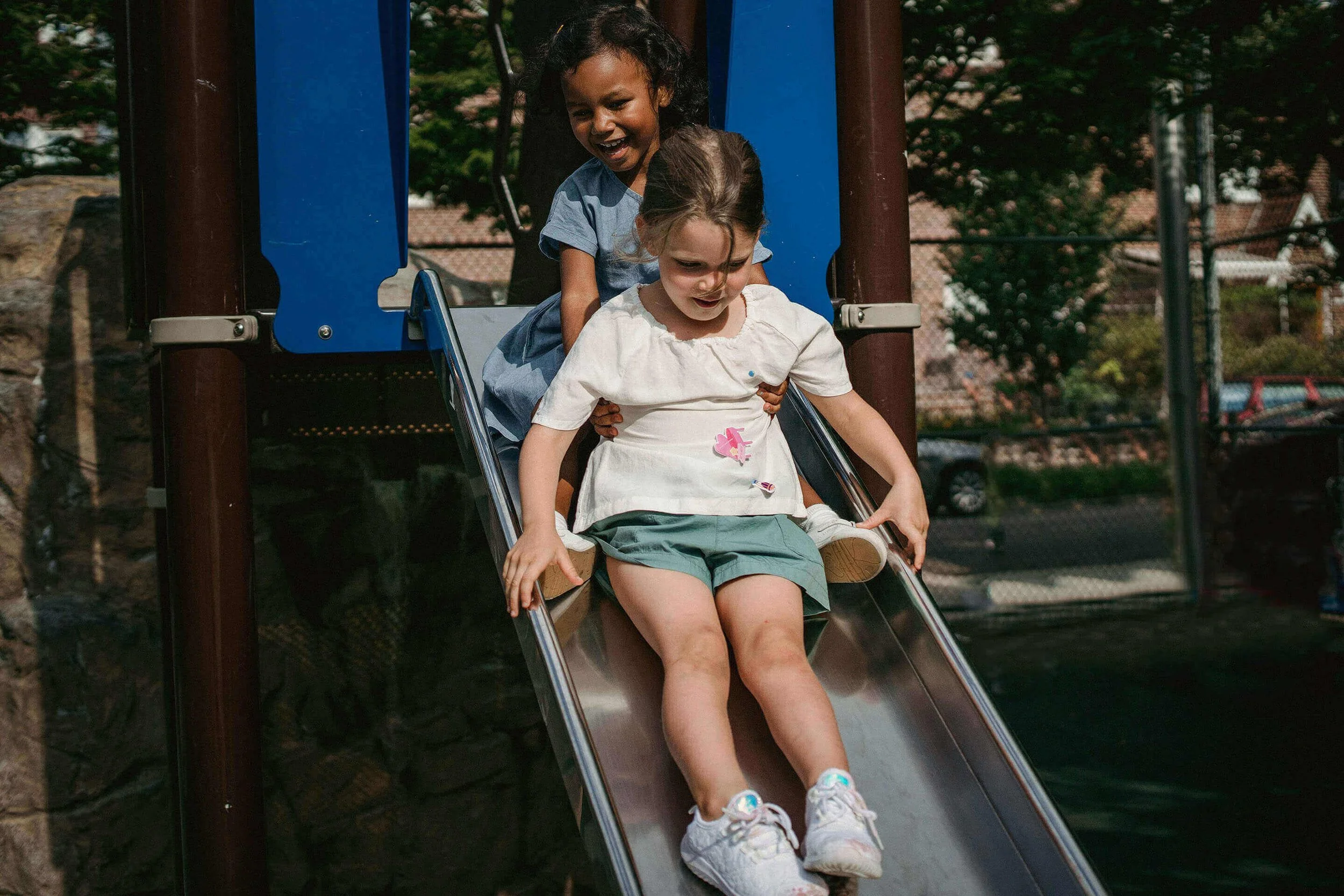Two young girls sitting at the top of a slide at a playground, smiling and enjoying themselves.
