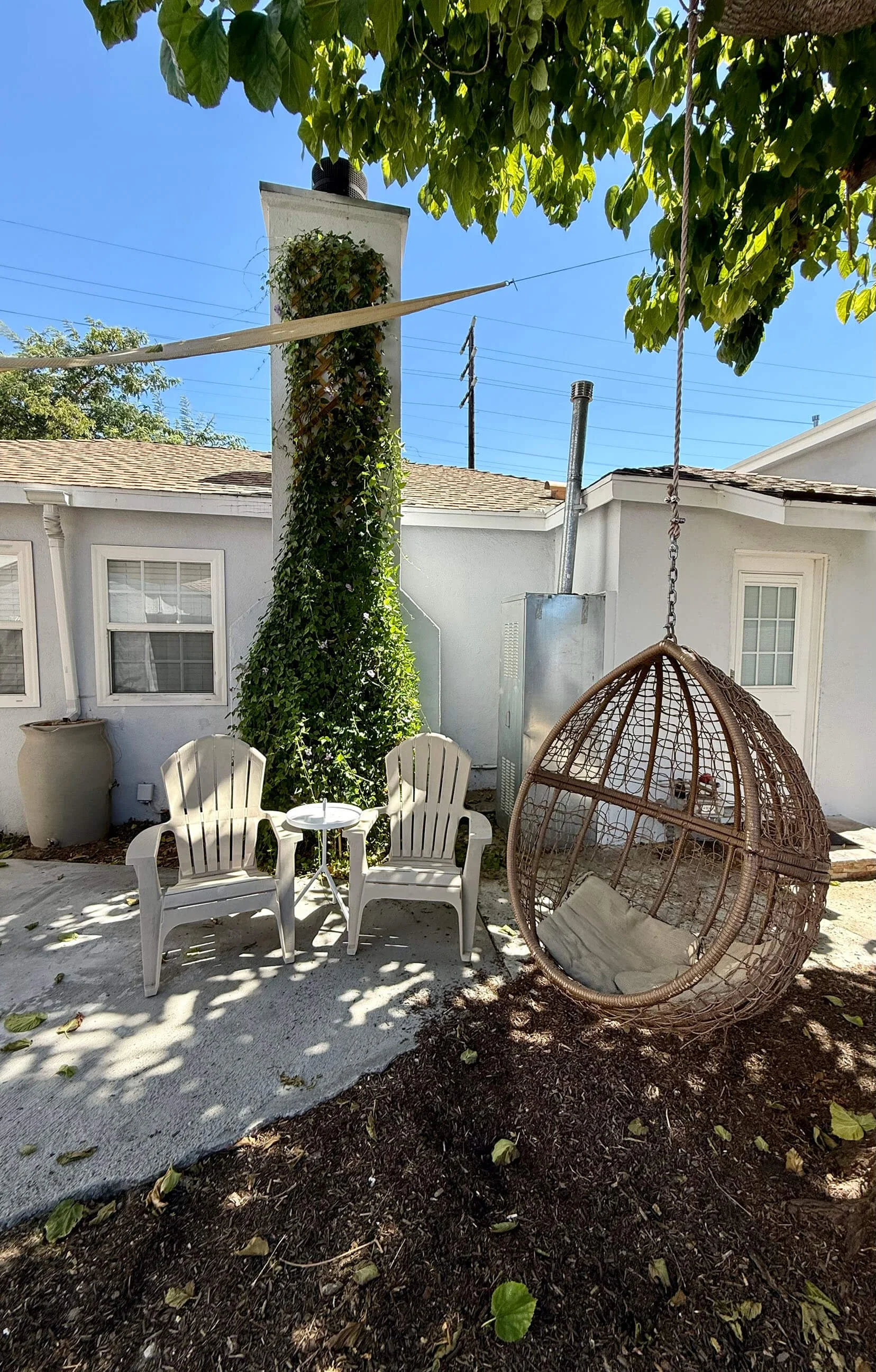 Residence 5645 & 5645 ½ Auckland Ave. A backyard patio with two white plastic chairs, a small table, a wicker hanging chair, and a large potted plant. There is a tall chimney and a leafy tree over the patio, with clear blue sky overhead.