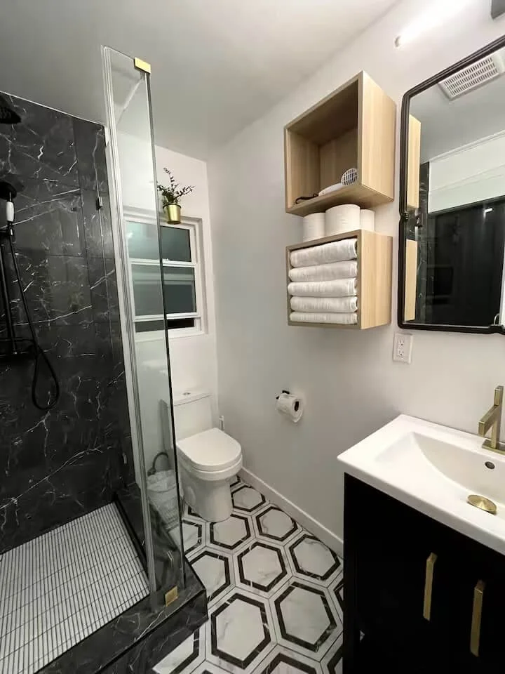 Modern white bathroom with black and white hexagonal tile flooring, black vanity with gold handles, glass-enclosed shower with black marble tiles, toilet, wooden wall shelves with towels and toilet paper, small window, mirror, and a small potted plan