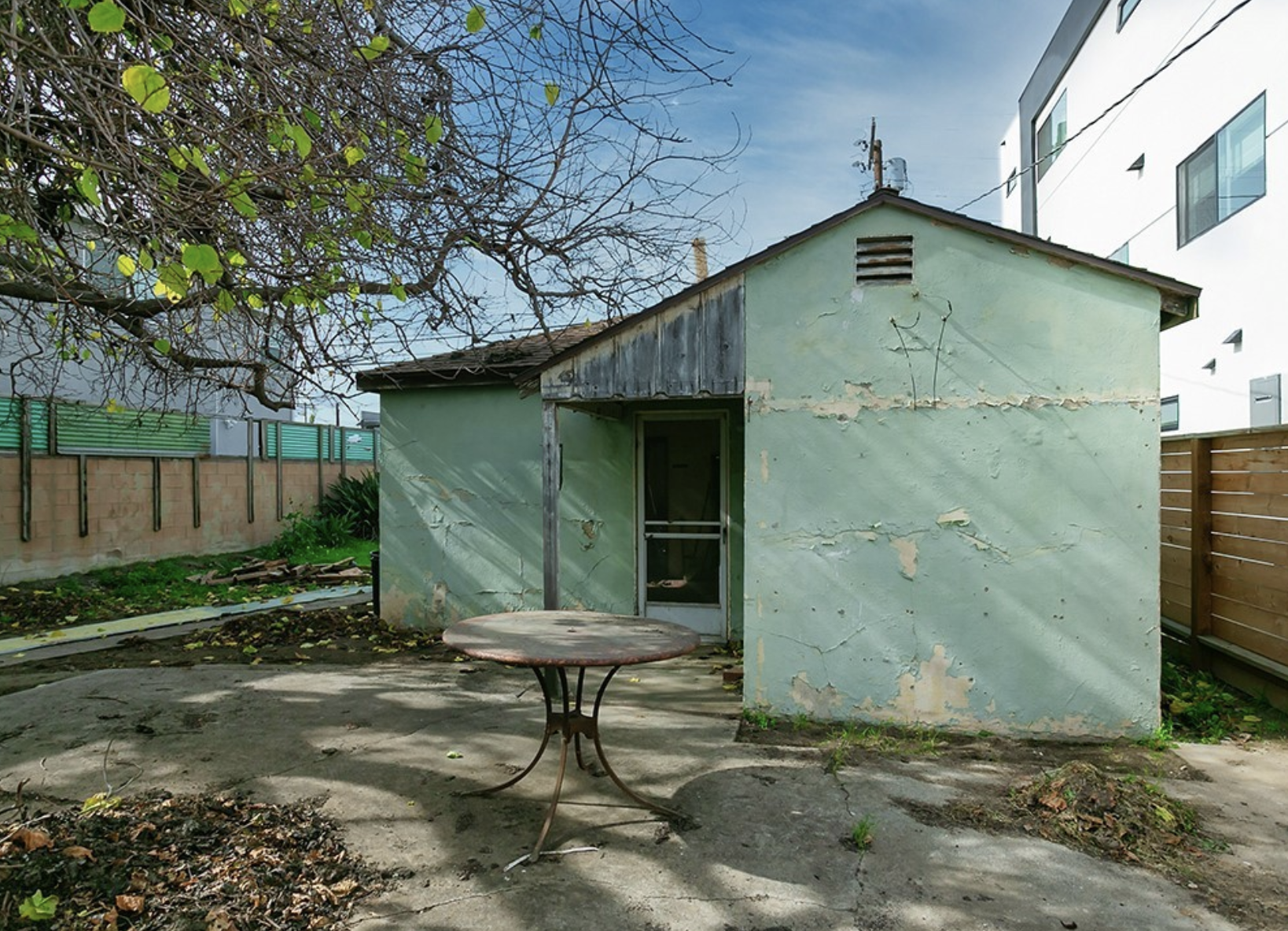 A small, abandoned building with peeling green paint and a partially covered doorway. There is a round, rusty metal table in front, with a tree casting shadows overhead. The background includes a wooden fence, some green plants, and a modern white bu