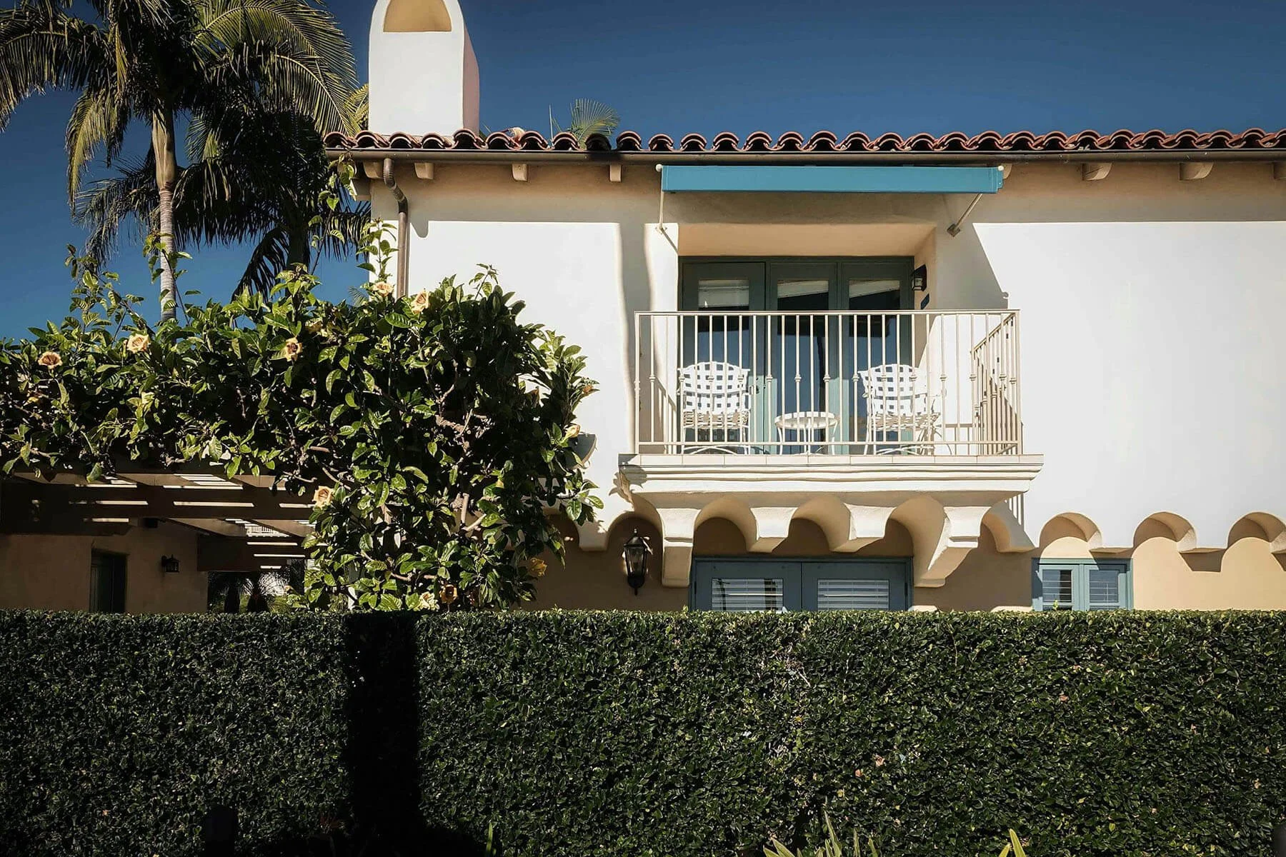The image depicts a two-story house with a balcony on the second floor, furnished with two chairs and a small table. The house has a red-tiled roof, white walls, and blue shutters. There is a large palm tree to the left and bushes with flowers in front, all set against a clear blue sky.