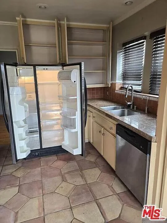 Empty kitchen with open refrigerator, beige cabinet drawers, a stainless steel dishwasher, sink with faucet, and window with blinds at residence 3016 W 83rd St. in Inglewood California.