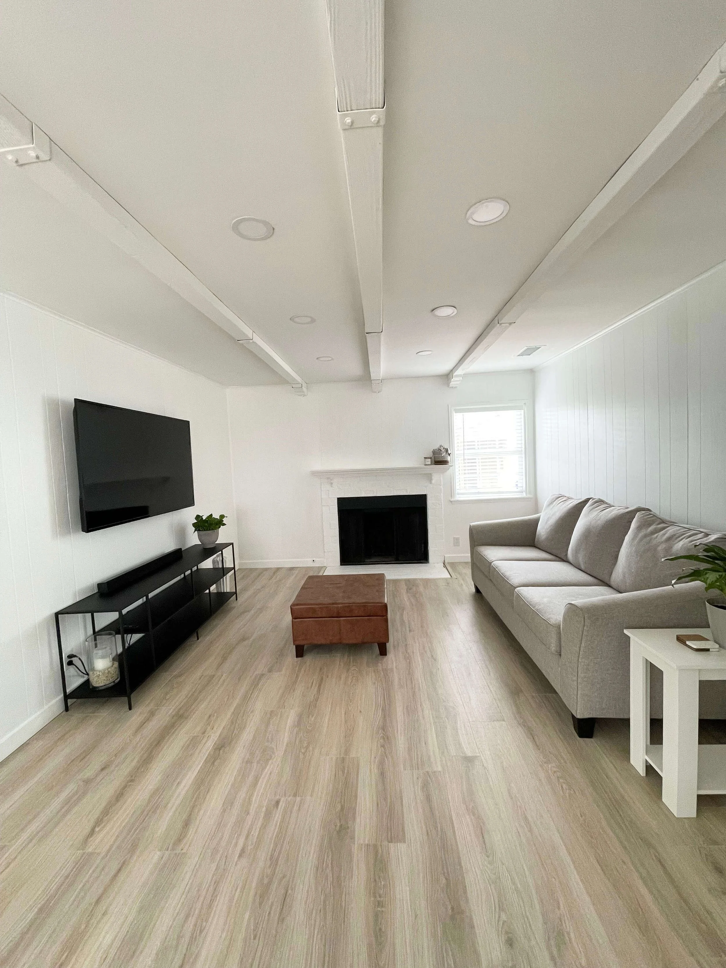 Residence 6605 & 6603 4th Ave. A living room with white walls, a beige sofa, a black TV mounted on the wall, a black media console, a small brown ottoman, a fireplace, a window with blinds, and light-colored wooden flooring.