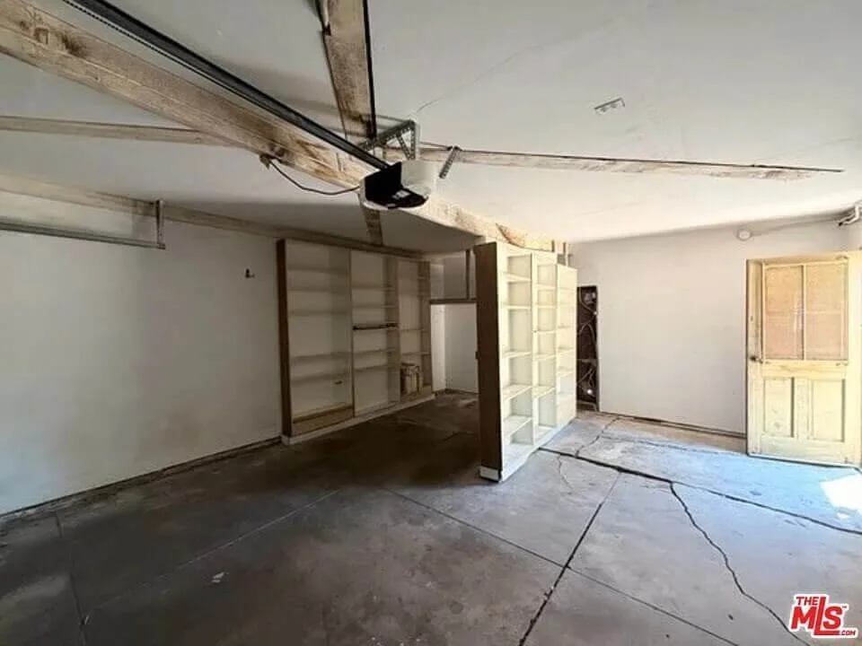 Empty room with cracked concrete floor, built-in shelves, a partially open window or door, exposed ceiling beam, and an overhead garage door mechanism at residence 3016 W 83rd St. in Inglewood California.