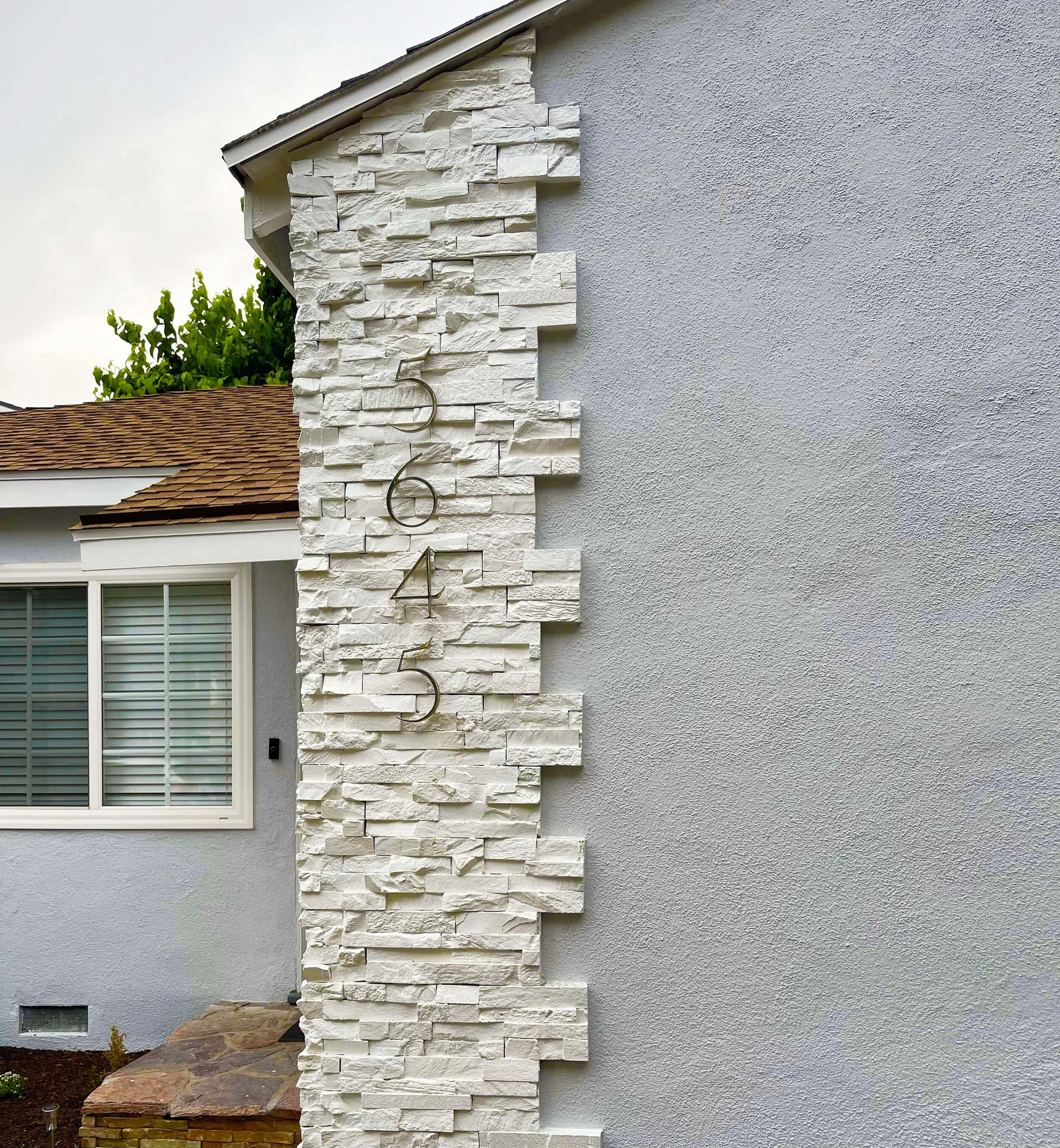 Close-up of a house exterior showing a section of white stone siding on the chimney and a smooth gray stucco wall. A window with white blinds and a small brick garden bed are also visible.