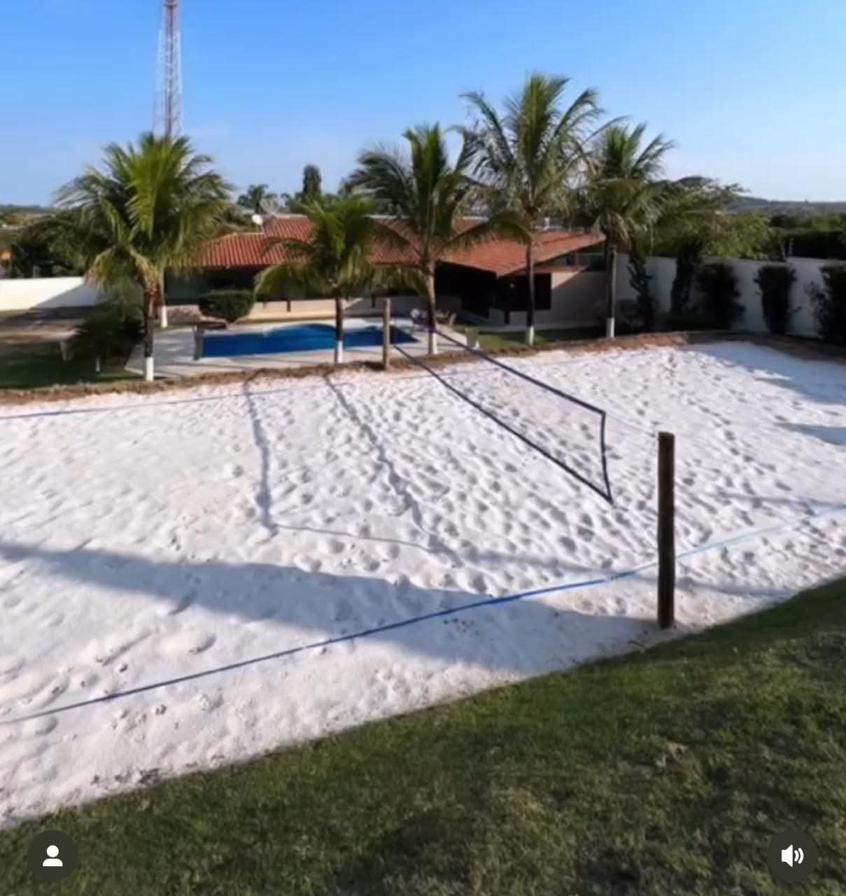 Quadra de vôlei de areia com rede, ao fundo uma piscina, árvores palmeiras e casas com telhado de barro, céu claro.