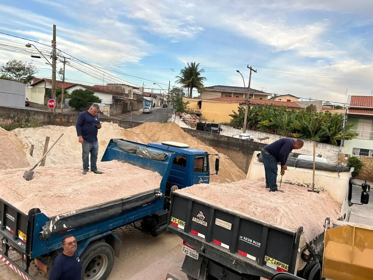 Homem trabalhando com areia em um caminhão de construção, com outro homem ao lado usando ferramenta e uma pessoa observando. Fundo com casas e árvores.