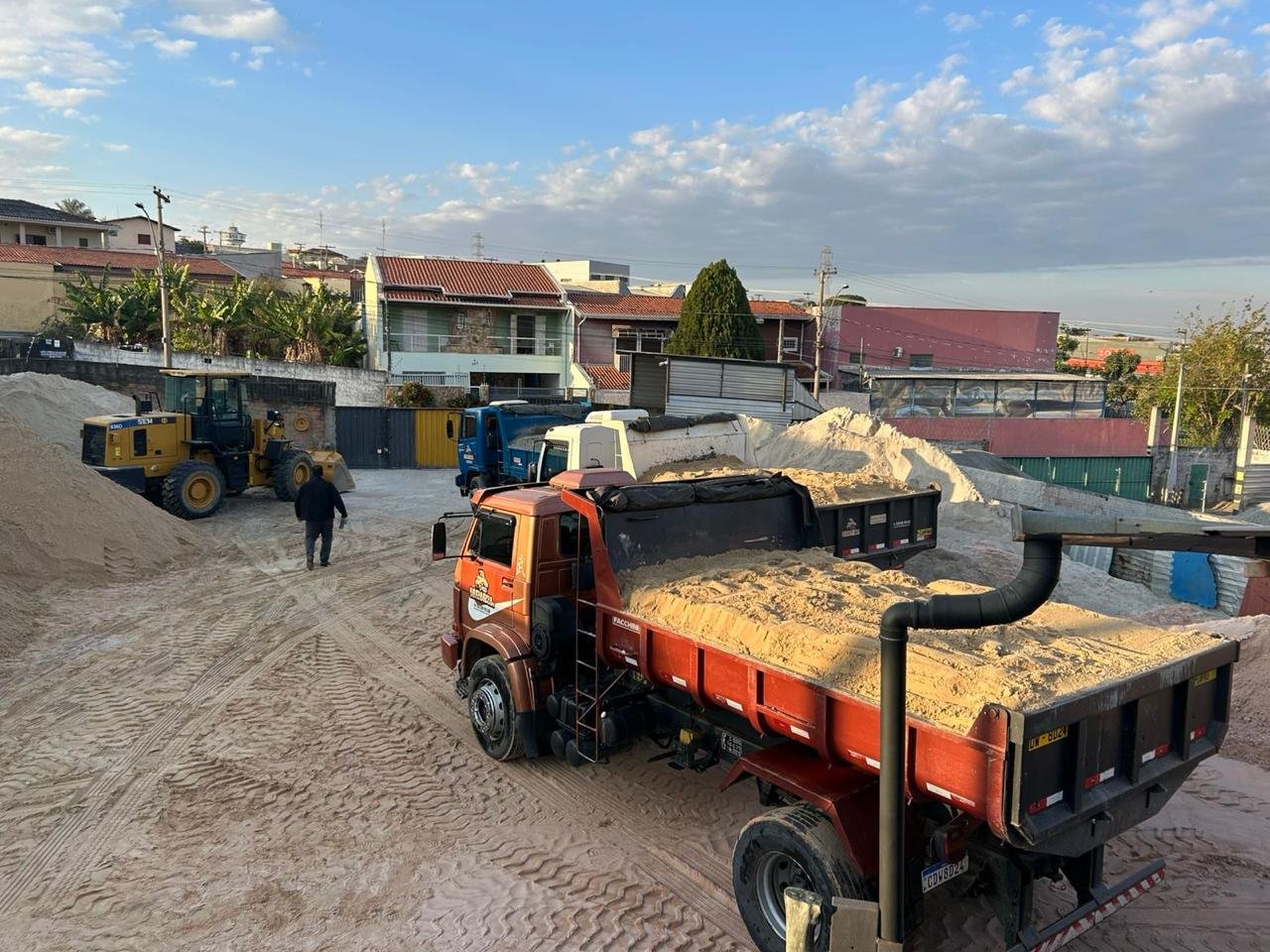 Construção de rua com caminhões de areia, uma escavadeira e um trabalhador. Casas e árvores ao fundo, céu com nuvens.