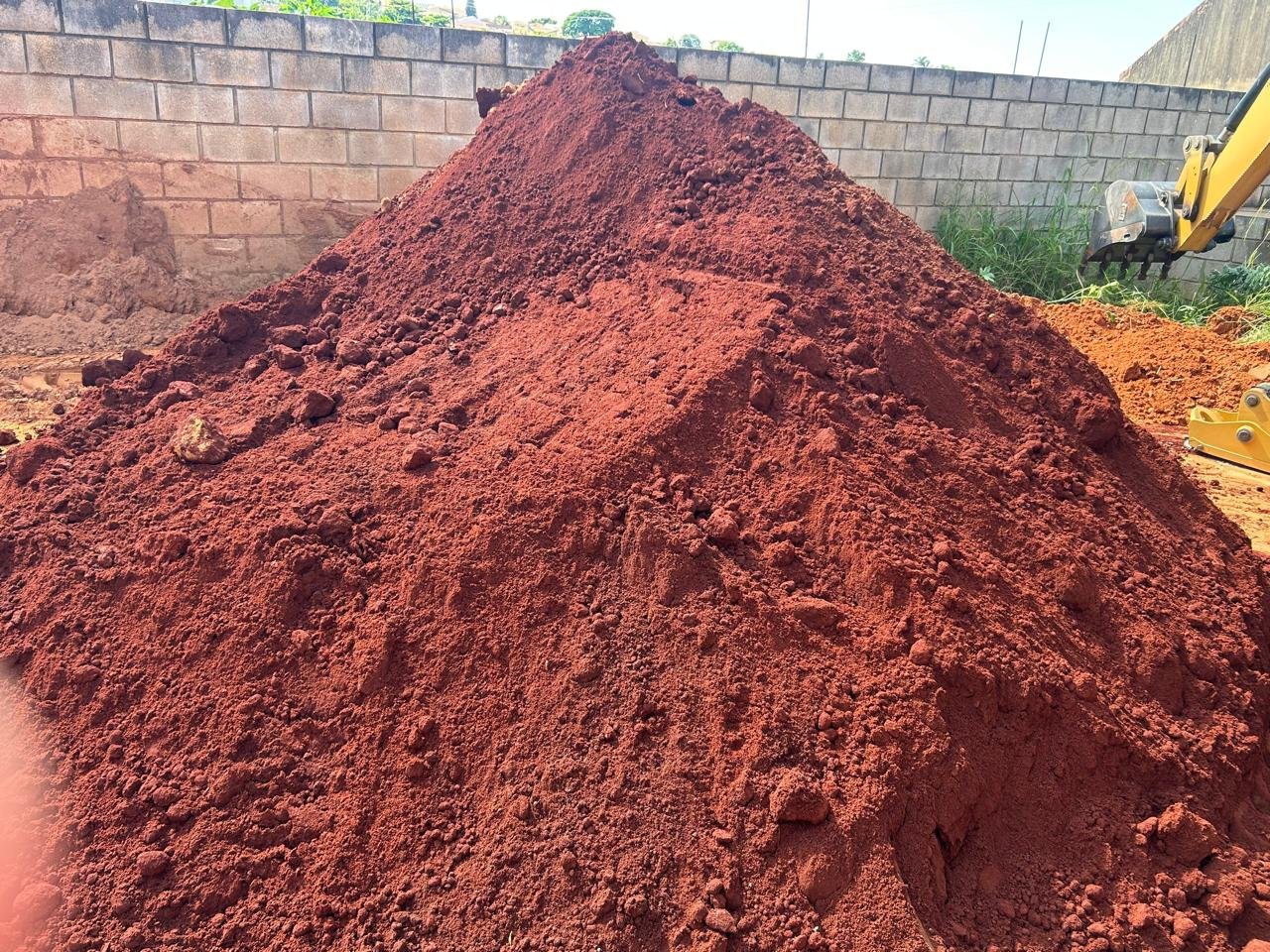 Pilha de terra vermelha em um canteiro de obras, com uma parede de blocos ao fundo e equipamento de escavação ao lado.