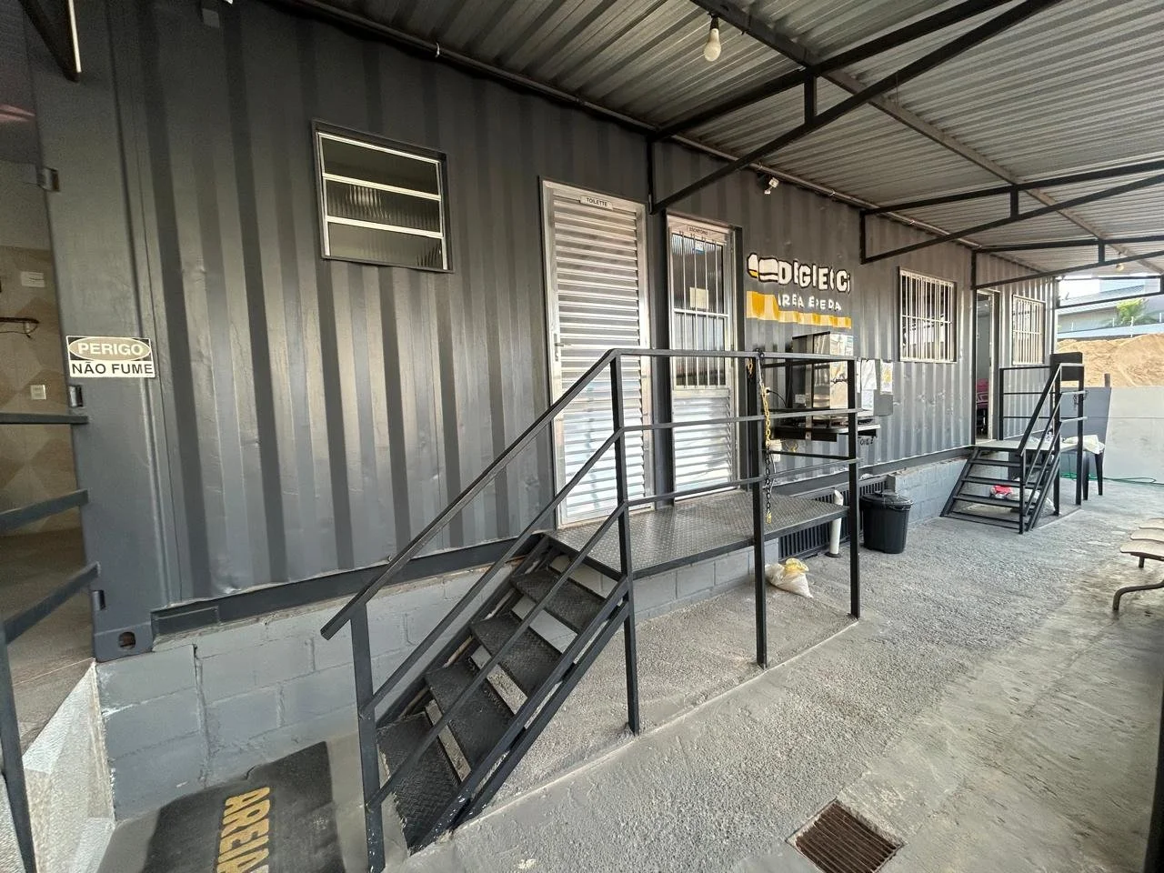 Área de entrada de uma estrutura metálica com escadas de acesso, sinalização de perigo, janelas com grades e um painel de venda de bilhetes.