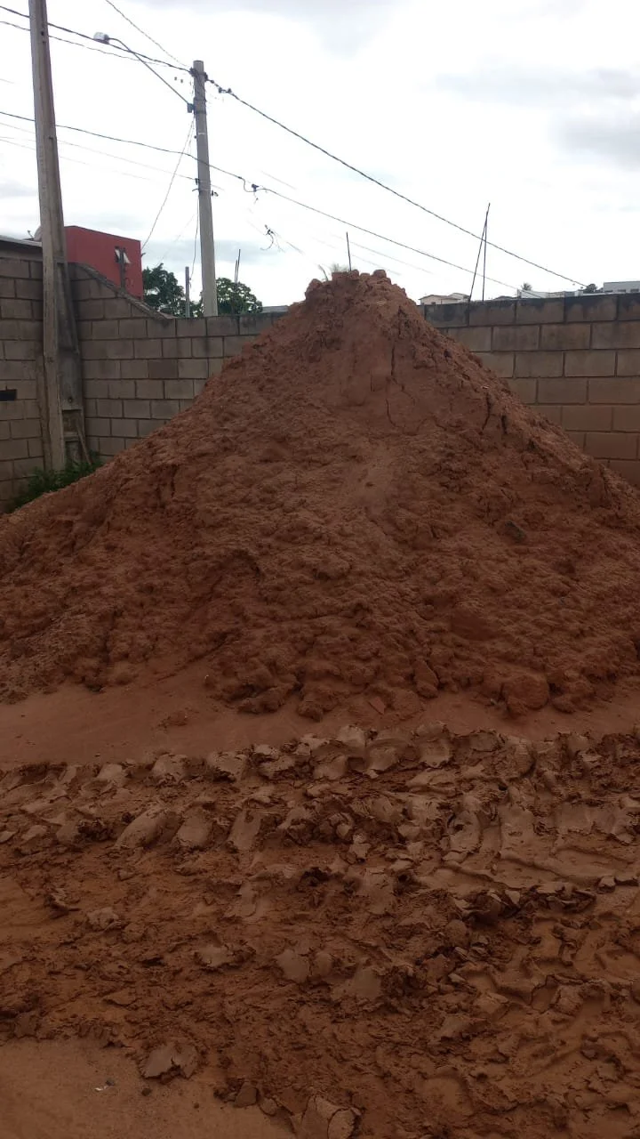 Montinho de terra vermelha em um quintal cercado por uma parede de blocos de concreto, com linhas de energia no fundo.