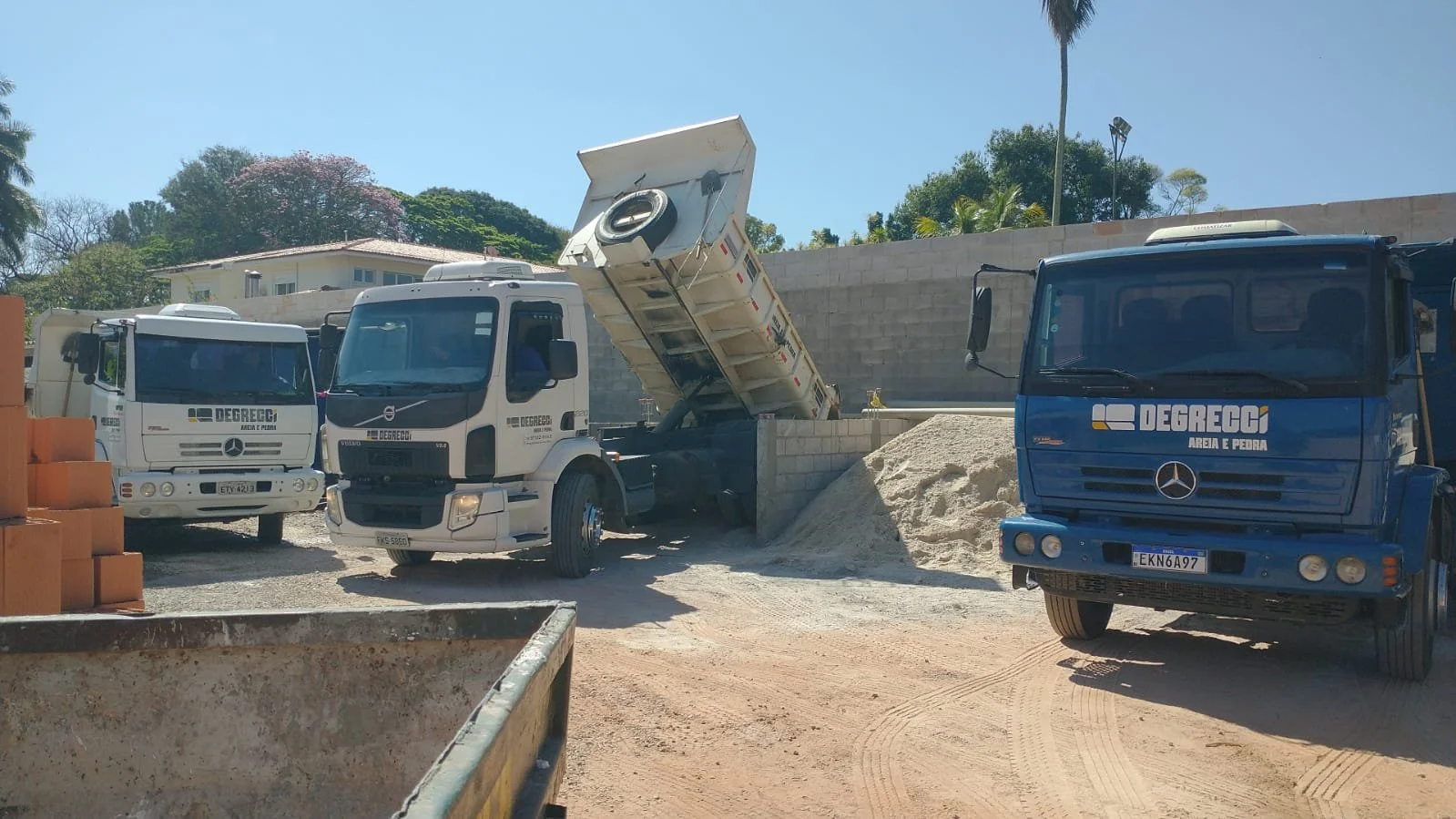 Caminhões de transporte de areia e pedra em um canteiro de obras com muro de bloco ao fundo