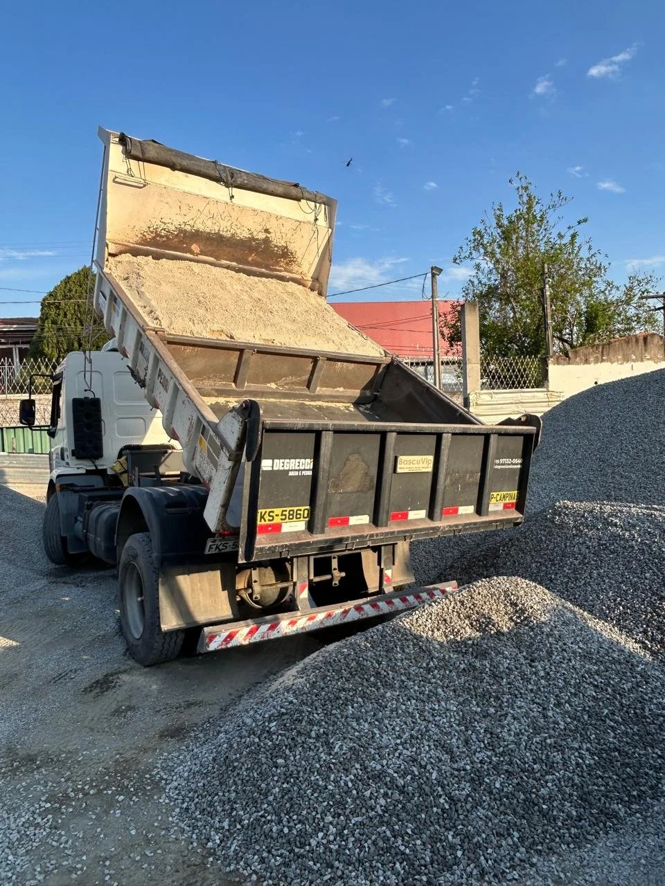 Caminhão de transporte de pedras cinza apiladas ao lado de uma pilha de brita, sob céu azul claro com poucas nuvens.