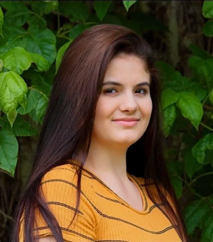 Lyndsey Ward: A young woman with long brown hair, wearing a yellow shirt with black stripes, standing outdoors in front of green leaves.