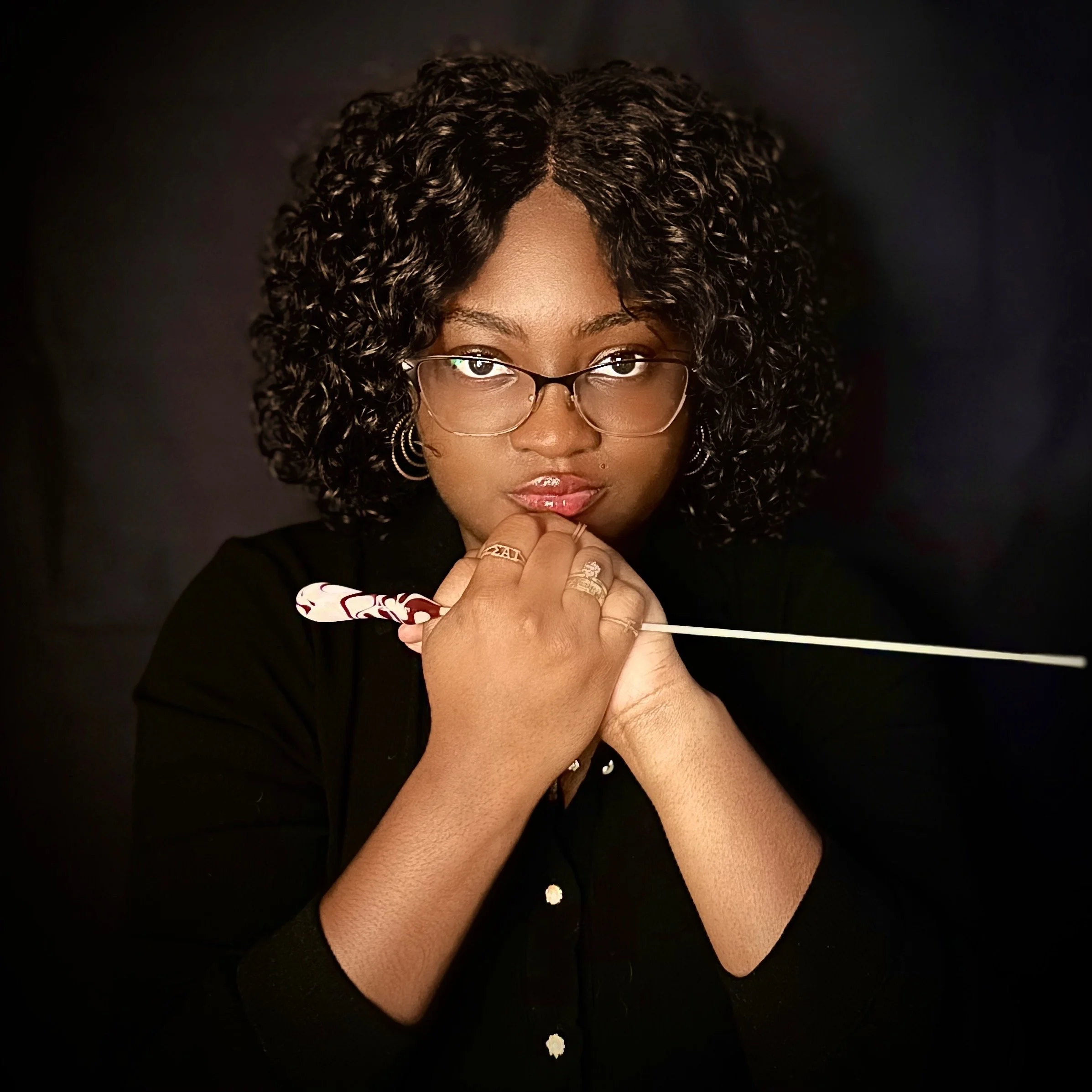 Jade Brazzell: A woman with curly hair, wearing glasses and earrings, looking directly at the camera with a serious expression, holding a conductor's baton against a dark background.
