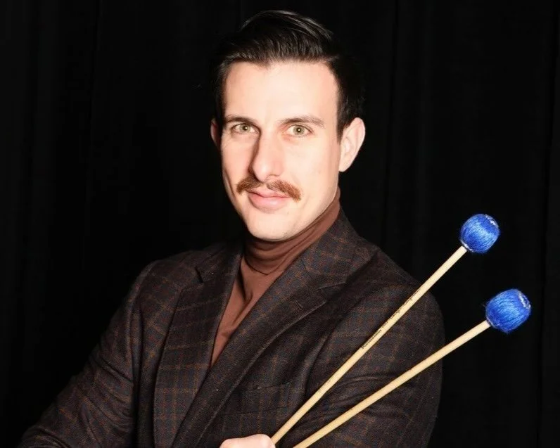 Michael Walling: Man with dark hair and mustache holding two blue mallets, wearing a brown shirt and plaid blazer, against a black background.