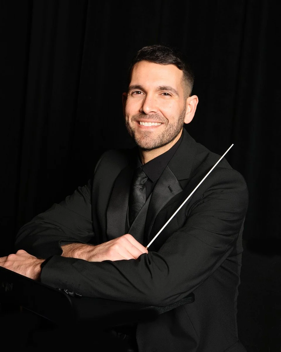 Zachary Johnson: A man with short dark hair, dressed in a black suit and tie, smiling while holding a conductor's baton, sitting  and against a dark backround