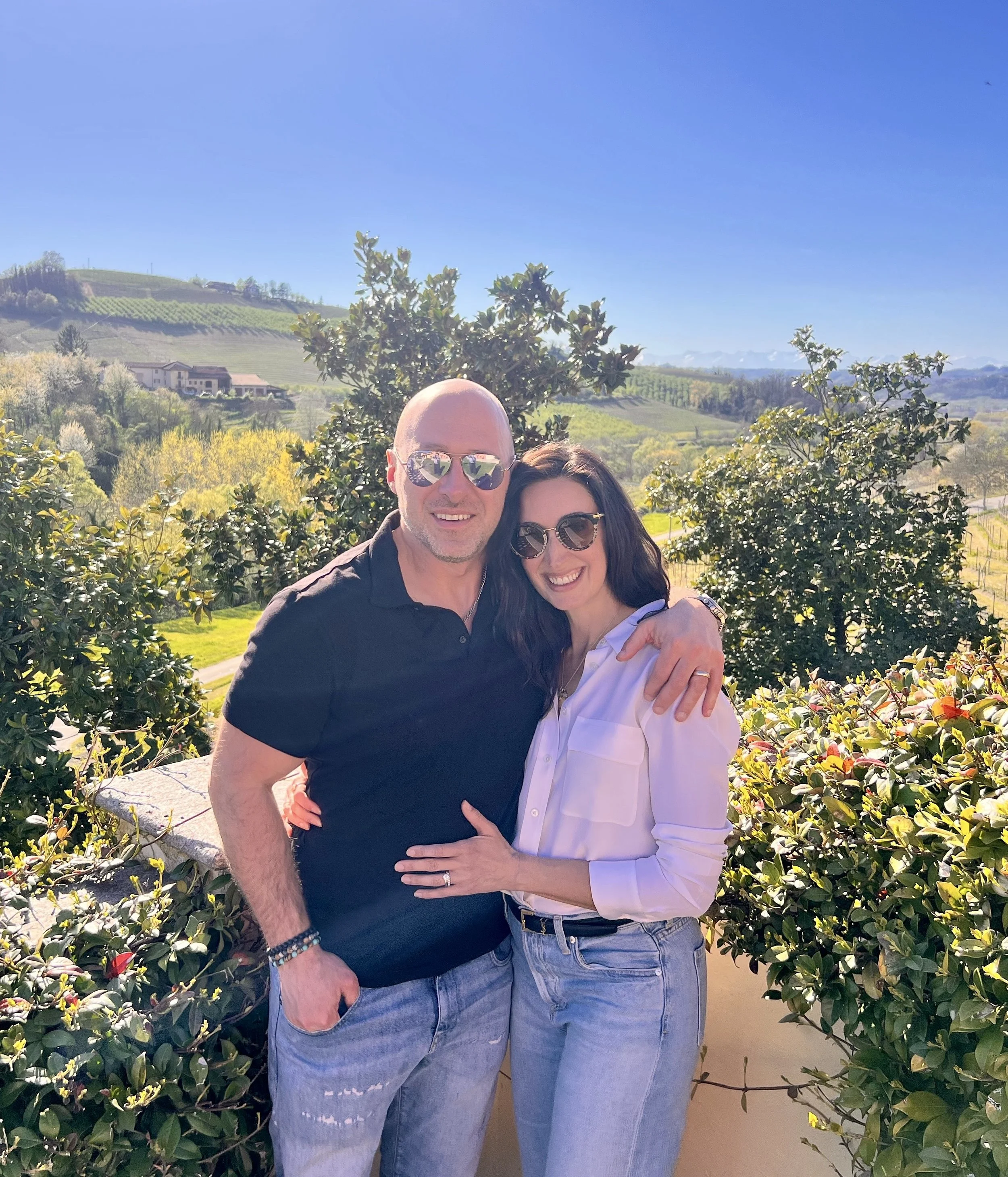 A man and a woman smiling and hugging outdoors among green bushes. The man is wearing sunglasses and a black shirt. The woman is wearing sunglasses, a white shirt, and has dark hair. Background shows rolling hills, trees, and a clear blue sky.