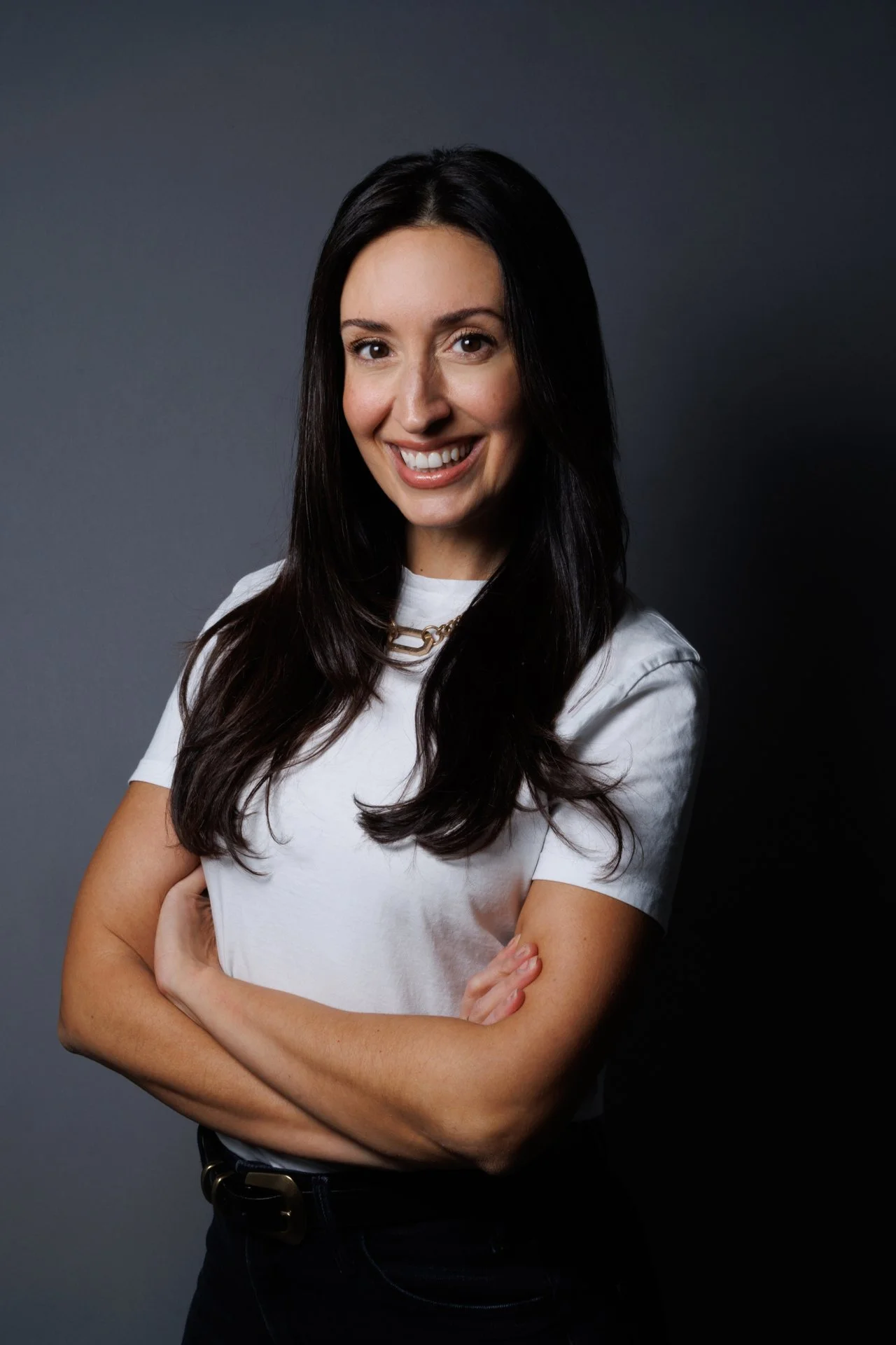 A woman with long dark hair, smiling, wearing a white t-shirt and a gold chain necklace, standing against a dark grey background.