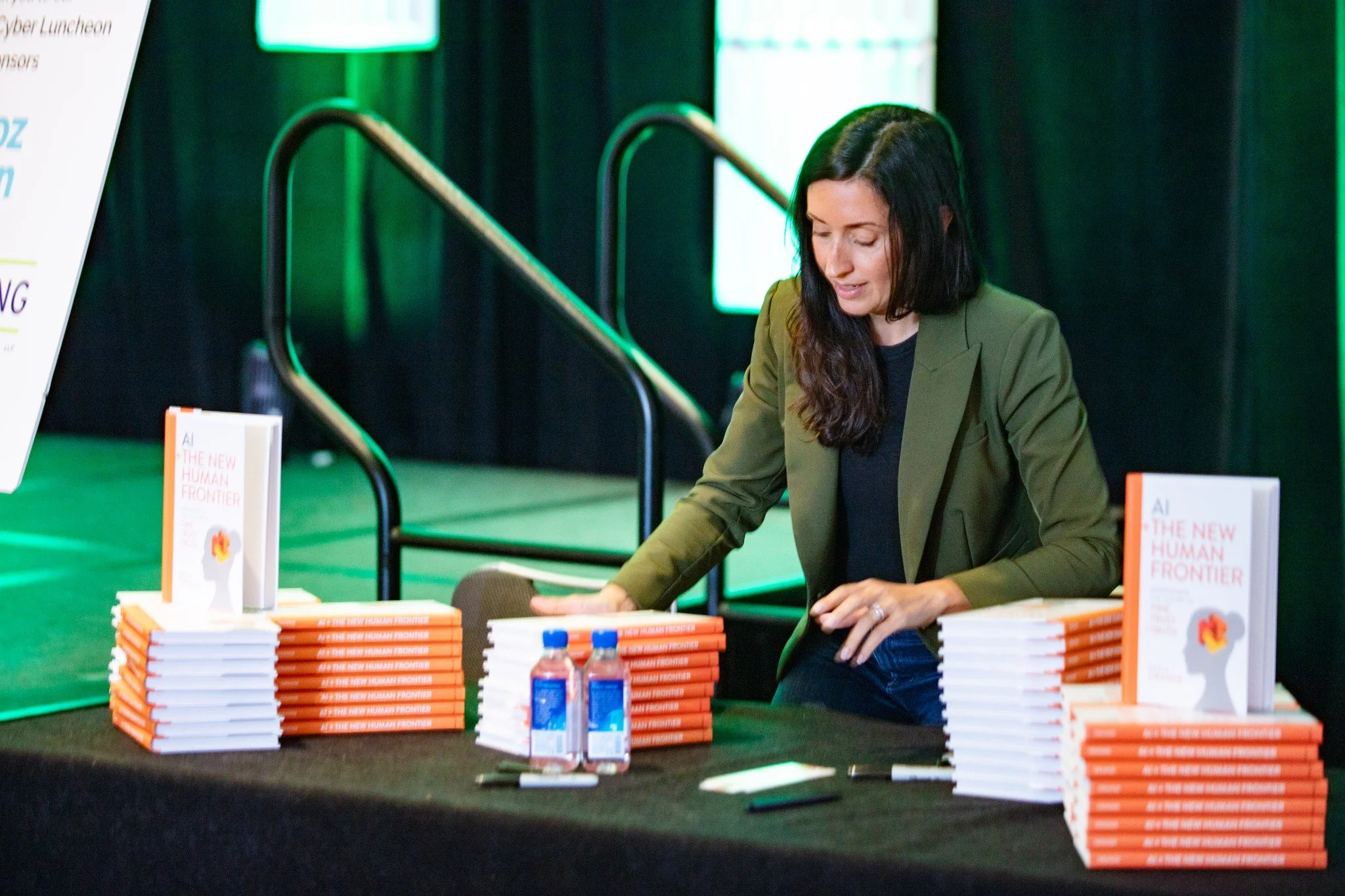 A woman with dark hair wearing a green blazer arranging stacks of books titled "AI: The New Human Frontier" at a book signing event, with promotional materials and water bottles on the table.