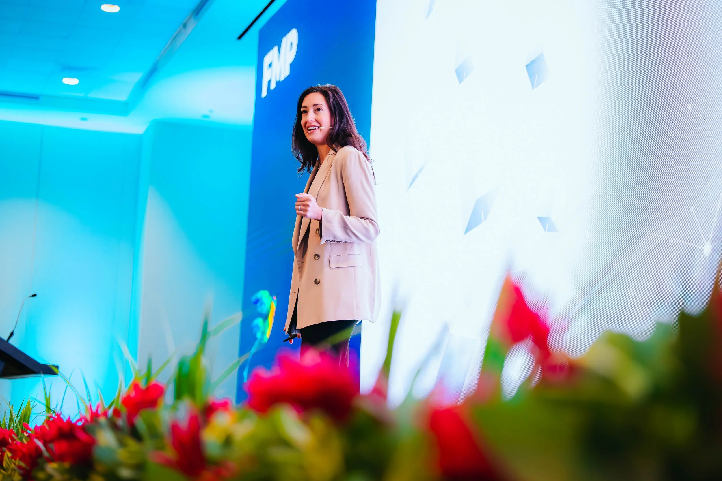 A woman in a beige blazer speaking at a conference with a blue backdrop displaying the Microsoft and Windows logos, seen from a low angle with red flowers in the foreground.