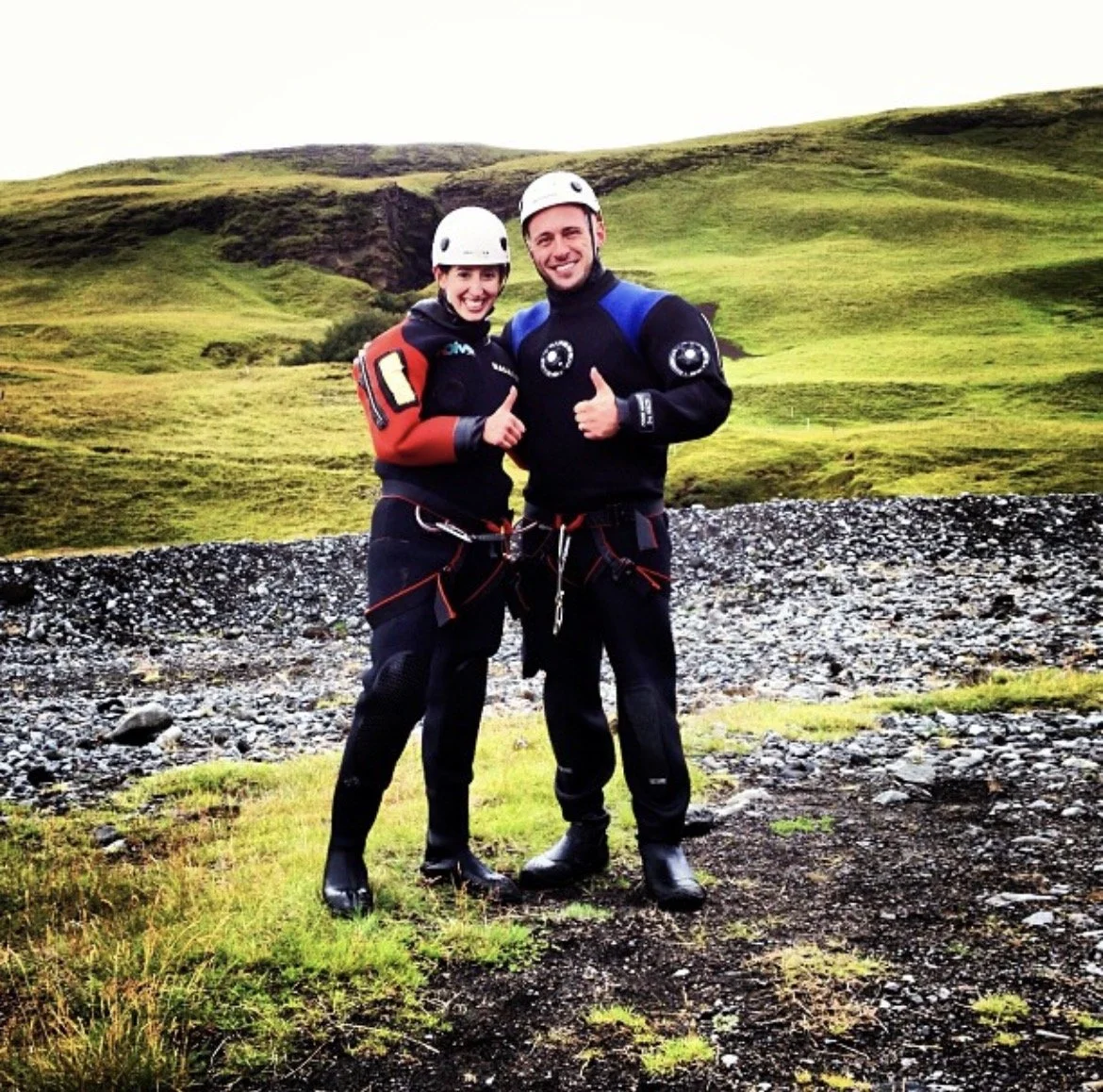 Two people wearing helmets and outdoor gear standing on rocky ground in a green, hilly landscape, giving thumbs up.