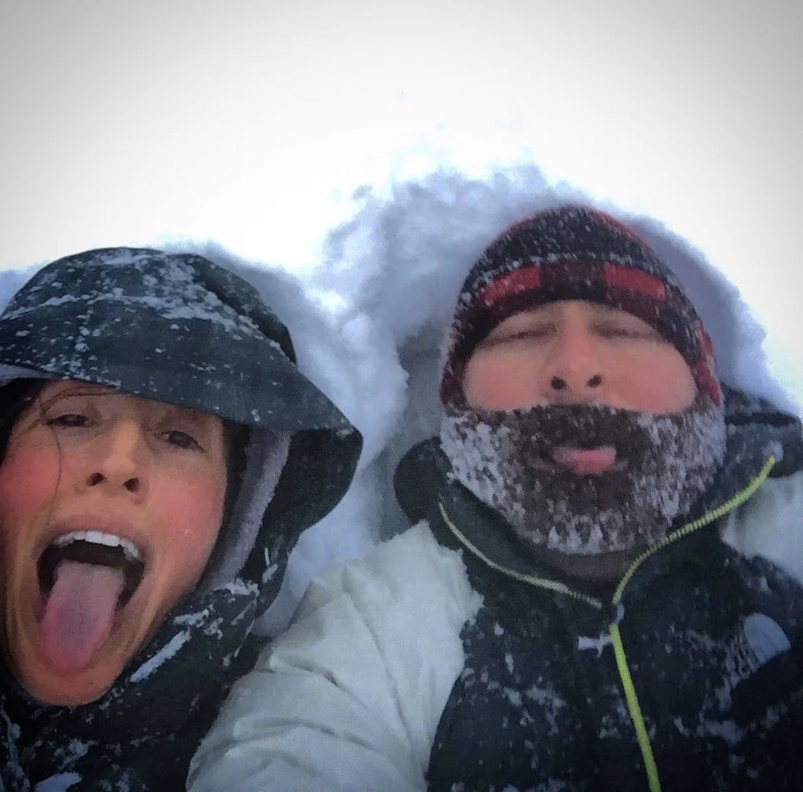 Two people bundled in winter clothing lying in snow, one with tongue out and the other with eyes closed, in a snowy outdoor environment.